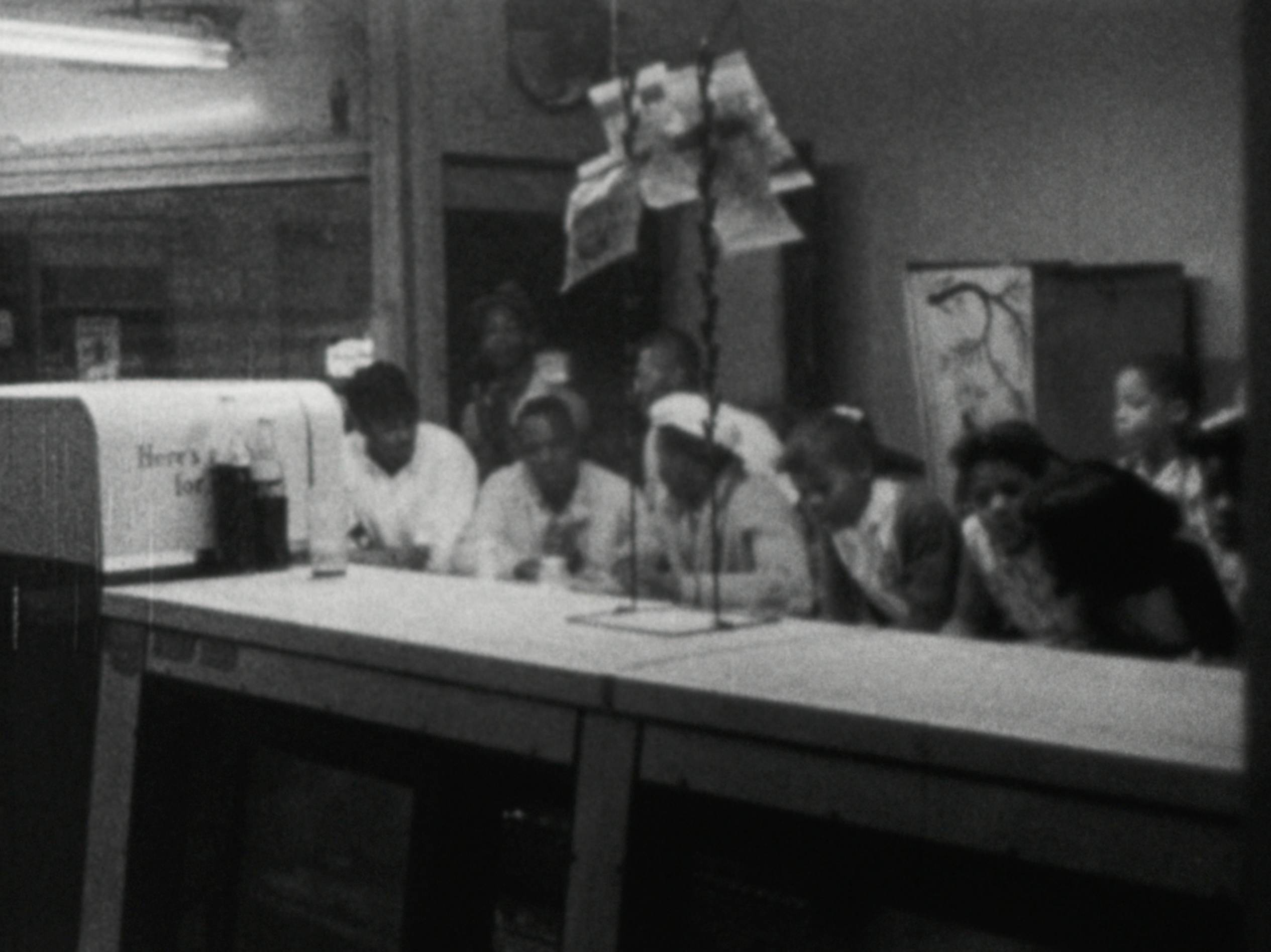 Photograph of a group of young people, mostly girls, seated at a long counter inside a diner or café. They are closely gathered, heads down, focused on writing or drawing. The foreground includes part of a counter with two glass soda bottles and a soft drink dispenser labeled “Here’s for.” A microphone with paper notes clipped above it hangs over the group. The background is dimly lit, with a framed sketch of a tree partially visible on the right.