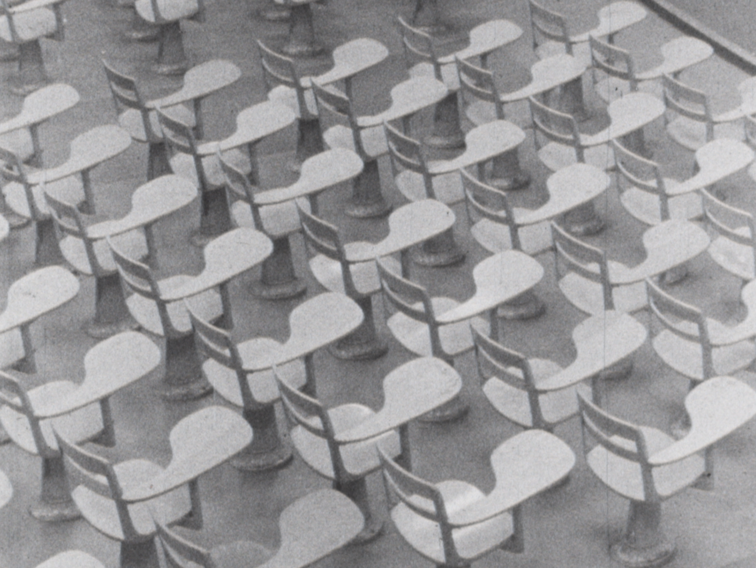 Black-and-white overhead view of an empty lecture hall filled with curved, molded-plastic chairs arranged in a staggered grid. Each chair has an attached writing surface shaped like an asymmetrical hourglass, supported by a single pedestal base. The repetitive layout creates a pattern of light and shadow across the bare floor.