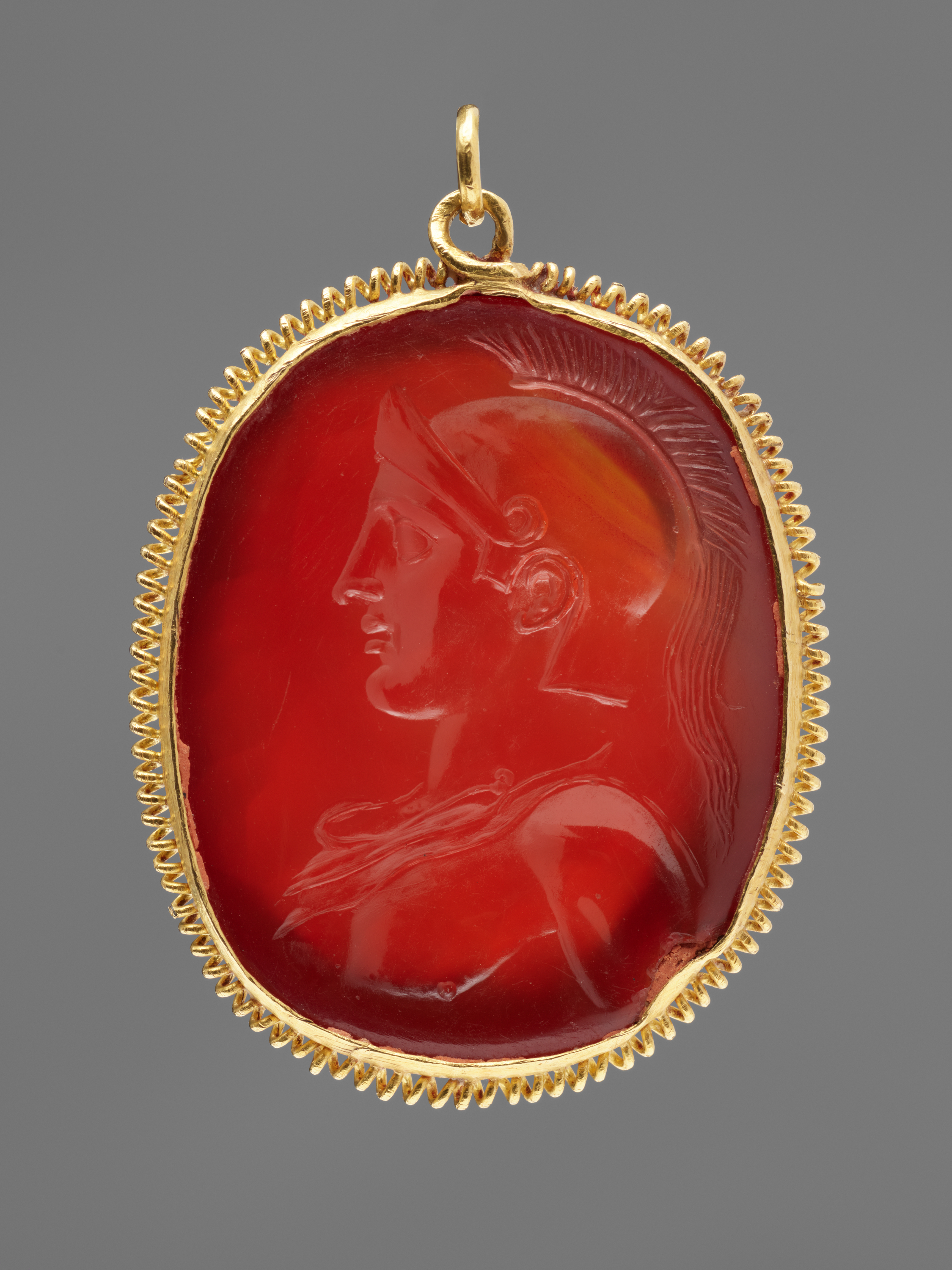A smooth red, oval gemstone in a gold setting features the incised carving of a clean shaven male figure in a plumed helmet.