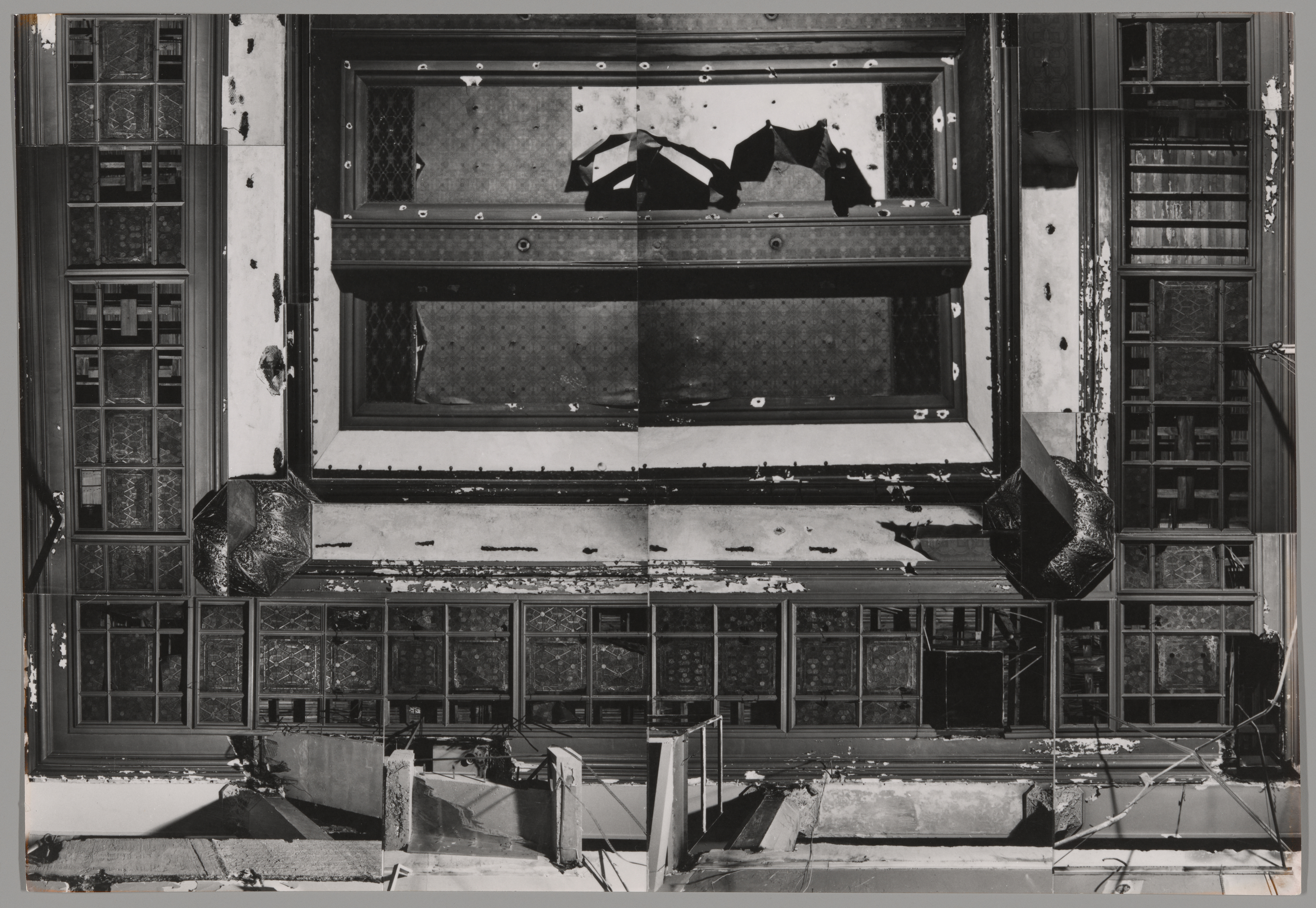 Black-and-white photograph of an ornate architectural ceiling seen from below, captured during or after significant damage. The ceiling features decorative moldings, coffered panels, and sections of glasswork, with peeling paint and visible structural wear. Large sections are broken or missing, allowing light to pass through. The image appears to be a composite of multiple photographs stitched together, emphasizing the symmetry and deterioration of the once-grand structure.