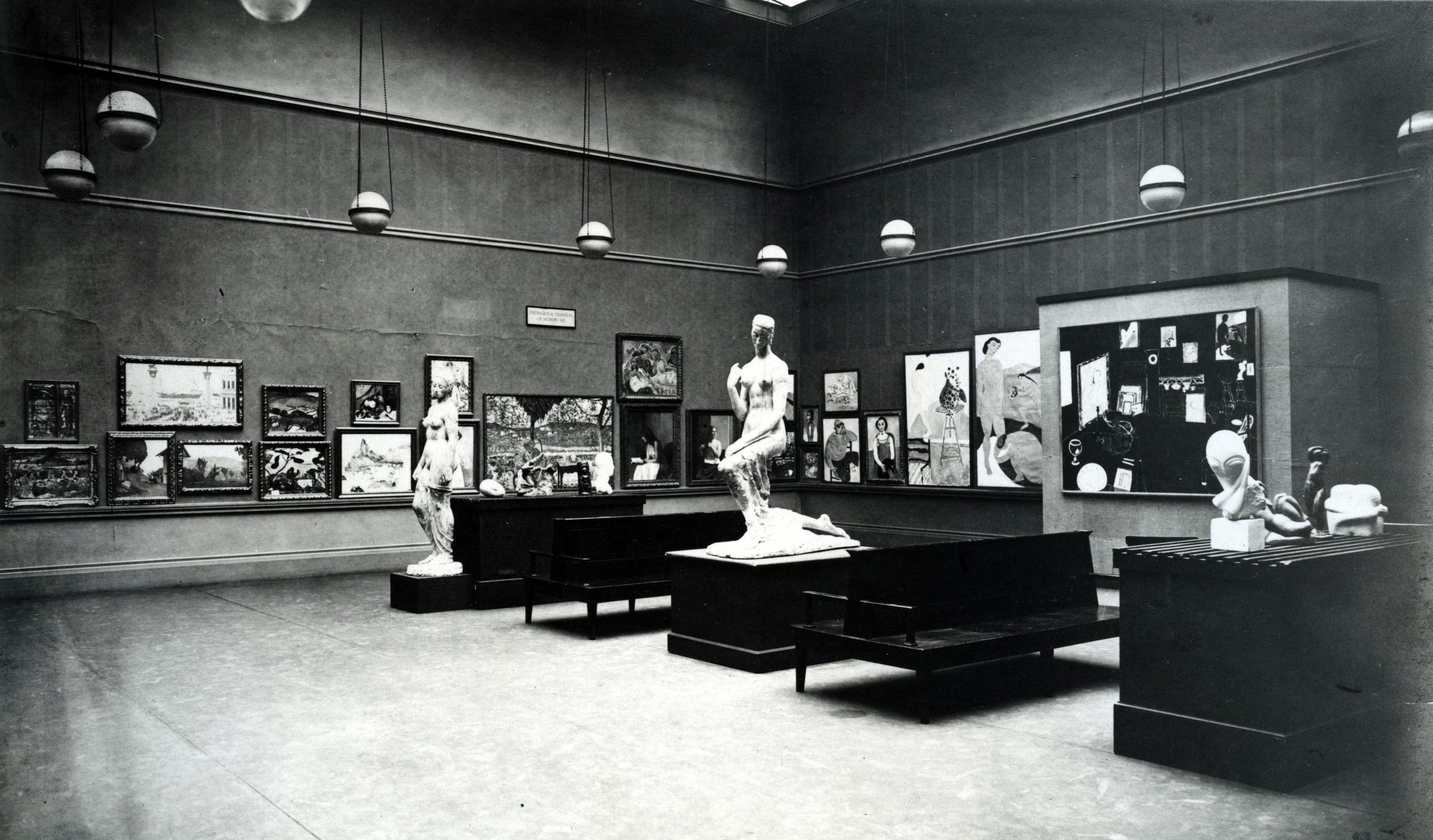 At the top, a high ceiling with several round white pendant lights hangs over gray walls. From left to center, a row of small to medium black-framed pictures lines the wall; in front stand two large white statues on pedestals—one upright and one kneeling—with long dark benches between them. On the right, more framed pictures appear, including one large panel filled with smaller images, and a raised platform holds several small smooth white sculptures; the foreground shows open floor space and two dark benches.