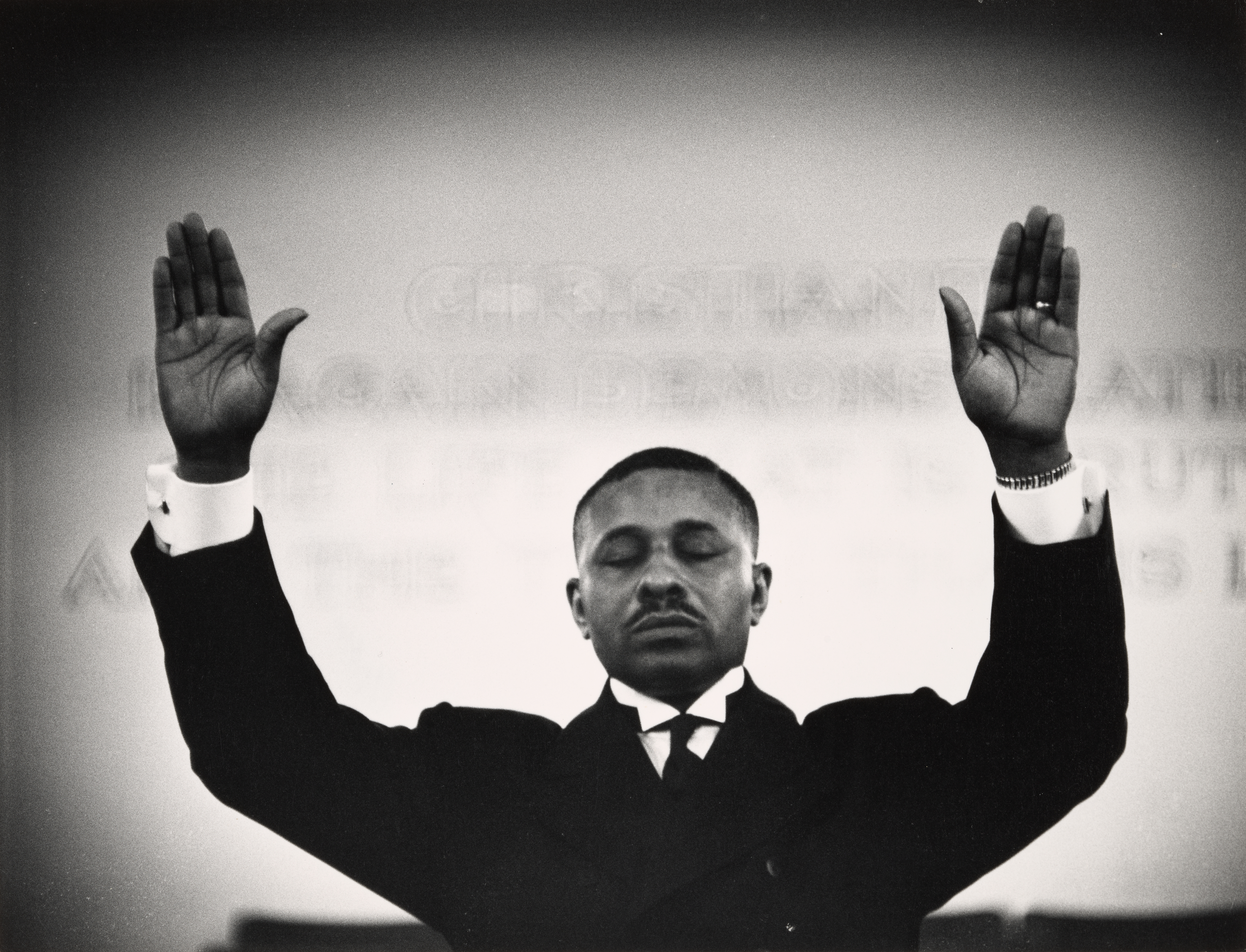A man in a dark suit and tie stands with his eyes closed and both arms raised, palms open and facing forward in a gesture of prayer or spiritual invocation. He wears a wristwatch on his right arm and cufflinks on his white shirt cuffs. The background is softly lit, with faint text visible on the wall behind him, suggesting a religious or church setting. The composition centers on the man’s serene expression and symmetrical pose.