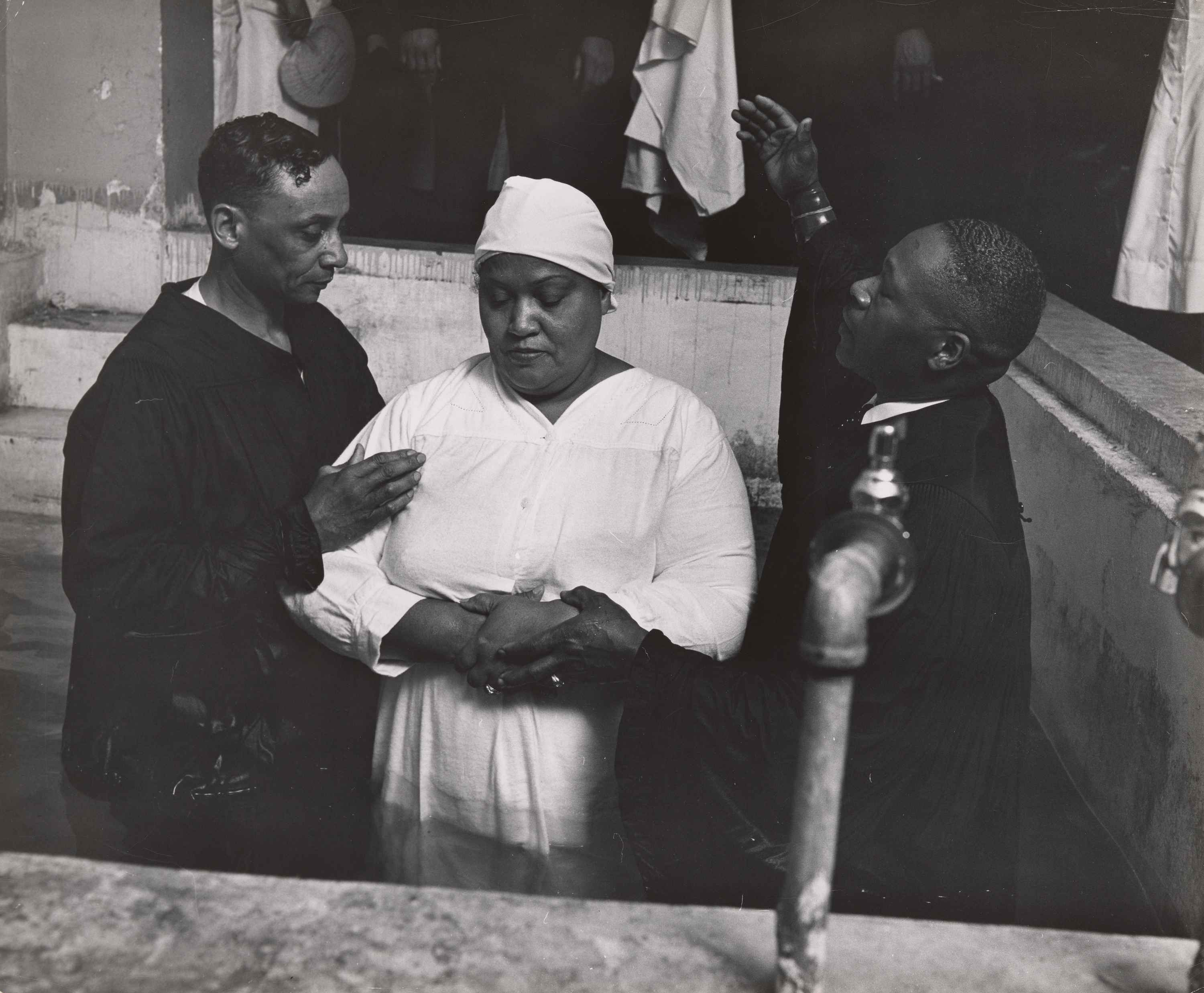 Three adults stand waist-deep in a baptismal pool inside a church. A woman in the center wears a white robe and head covering, holding her hands clasped in front of her. On either side, two men in dark robes guide the ritual—one places his hand gently on her shoulder, while the other raises one arm in a gesture of blessing or prayer. A pipe juts into the foreground at the right, and figures in white robes stand in the shadows behind them on the steps. The mood is solemn and reverent.
