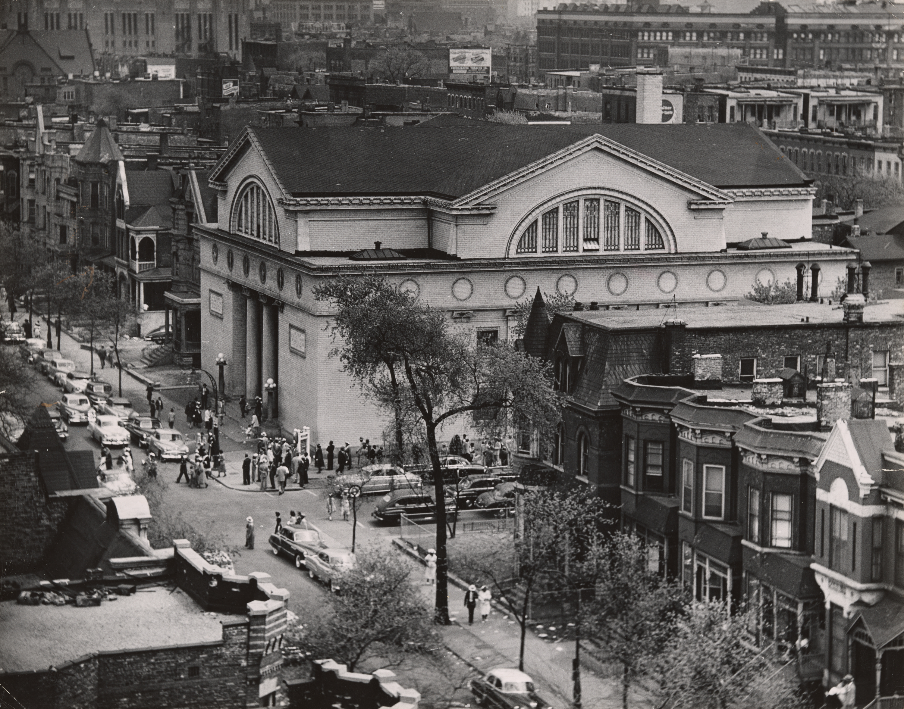 Aerial view of a large crowd gathered outside a neoclassical-style building with arched windows and a columned entrance, situated on a corner lot in a dense urban neighborhood. The scene includes several 1950s-era cars parked along tree-lined streets, with rows of brick residential buildings and businesses surrounding the area. People are seen walking on sidewalks and congregating near the building’s entrance, suggesting a public event or service. The photo captures the layered architecture and dense layout of the surrounding city blocks.