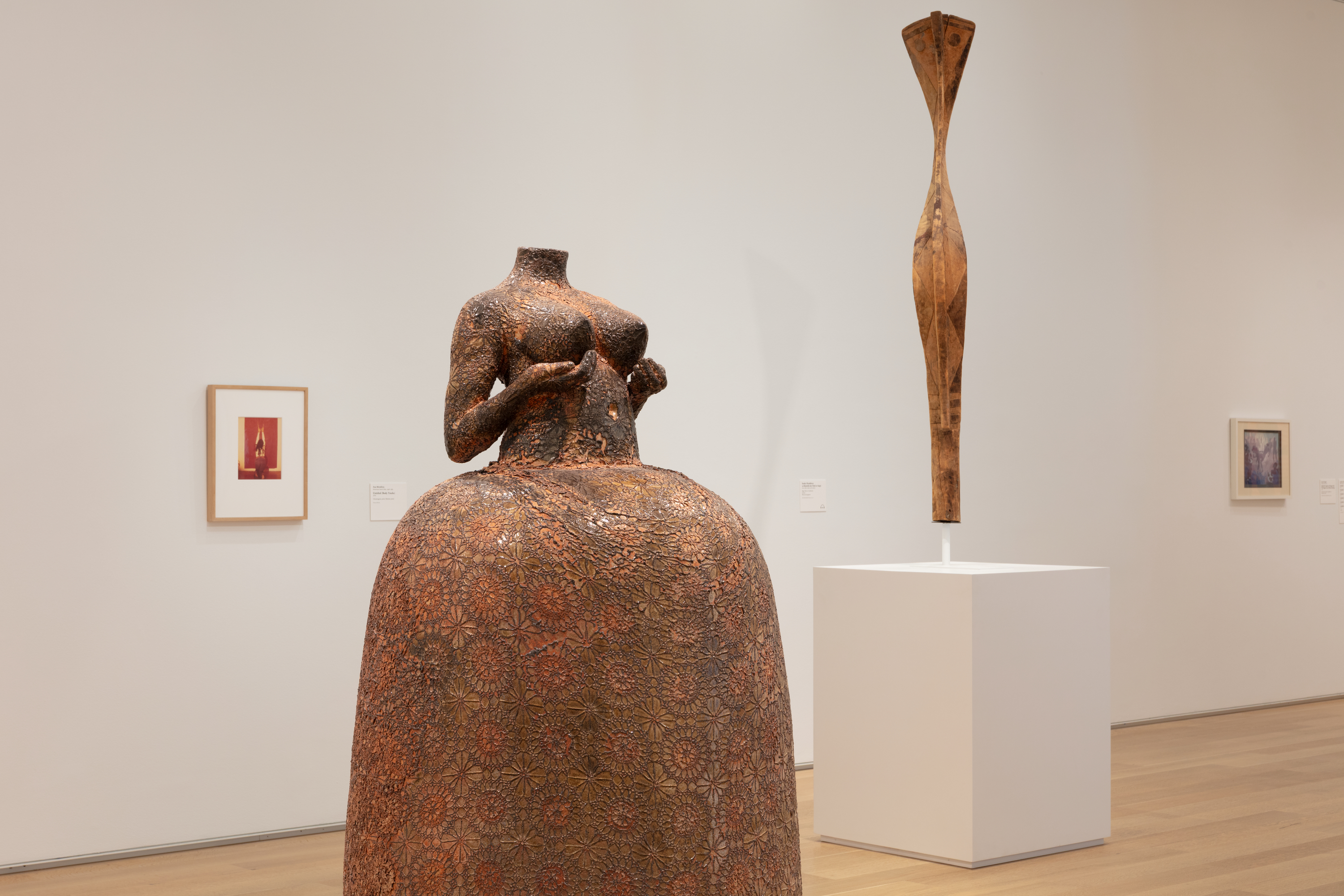 Photo in a white-walled art gallery shows a coppery-brown, life-size sculpture of a headless woman, naked from the waist up, hands nearly cupping the breasts, in a full, bell-shaped skirt. The sculpture is uniformly patterned with an imprinted floral motif. A small red photo is on the wall at left with a thin, spindly sculpture at right.