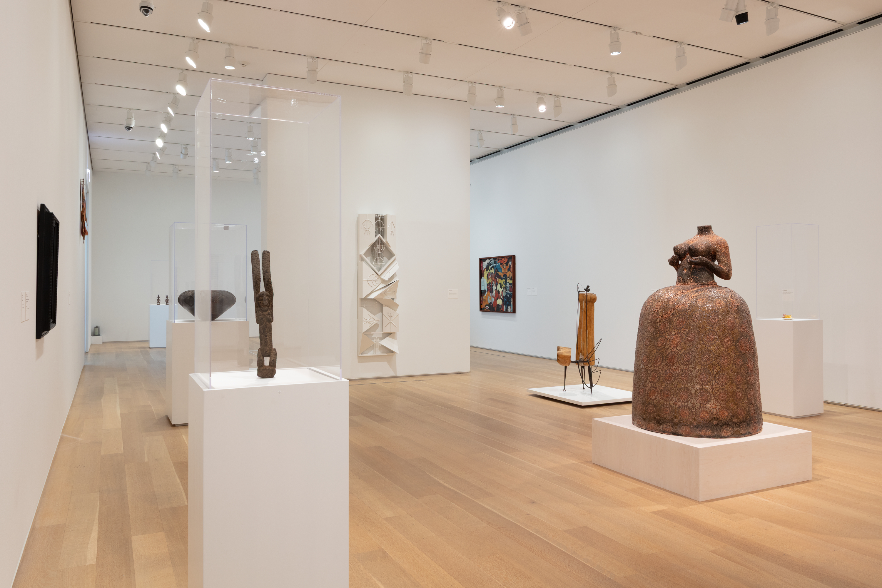 Photo of an art gallery with white walls, partitions and vitrines. At right is a large copper-colored sculpture of a headless woman, nude from the waist up, with a full skirt. At left, in a vitrine, is a sculpture of a humanoid figure with arms raised. Numerous other artworks of various mediums are evenly spaced throughout the gallery.