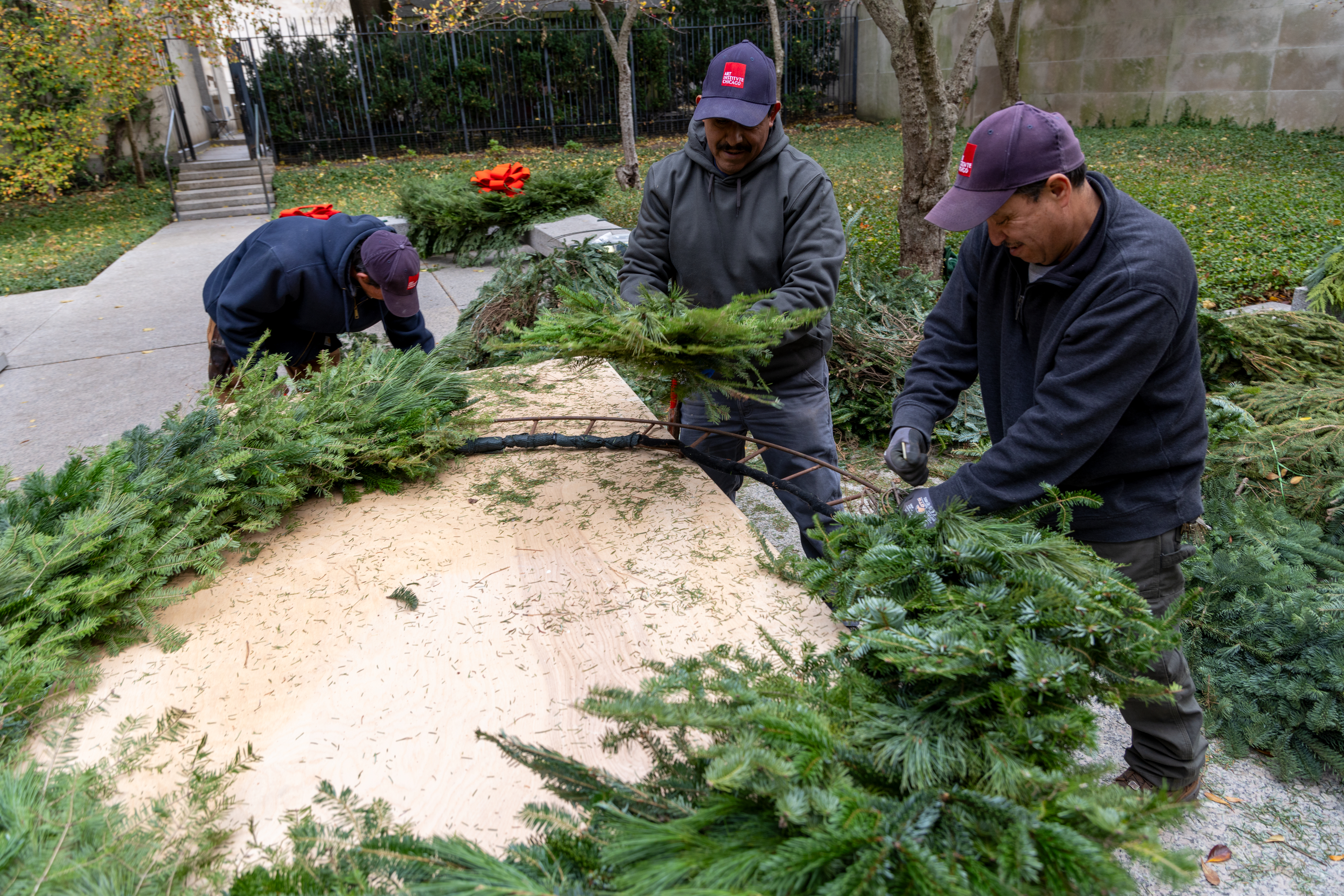 In a photo, three men work at an iron form, attaching evergreen branches.