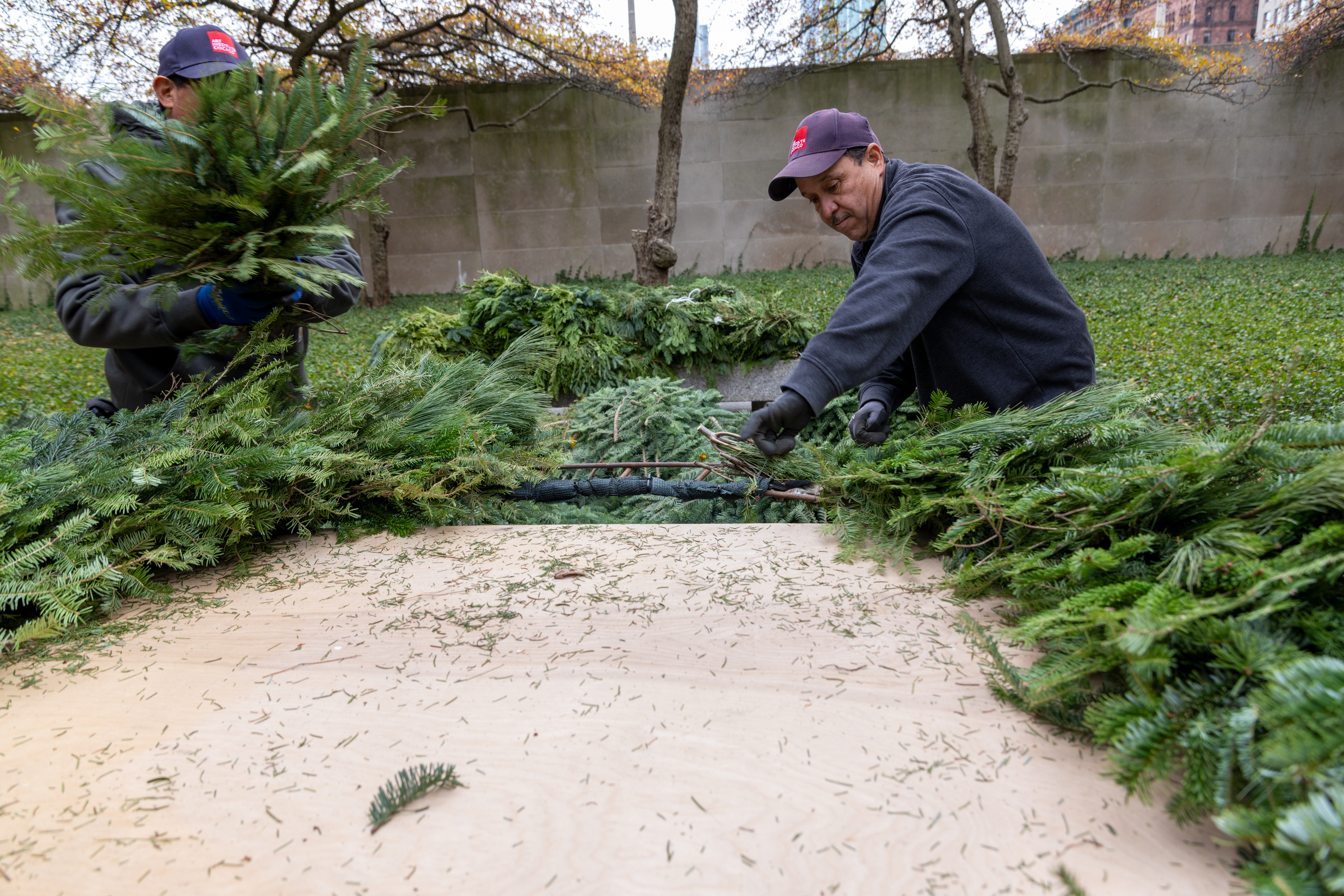 Photo shows a medium-skinned man in a blue sweatshirt working to attach evergreen branches to an iron form.