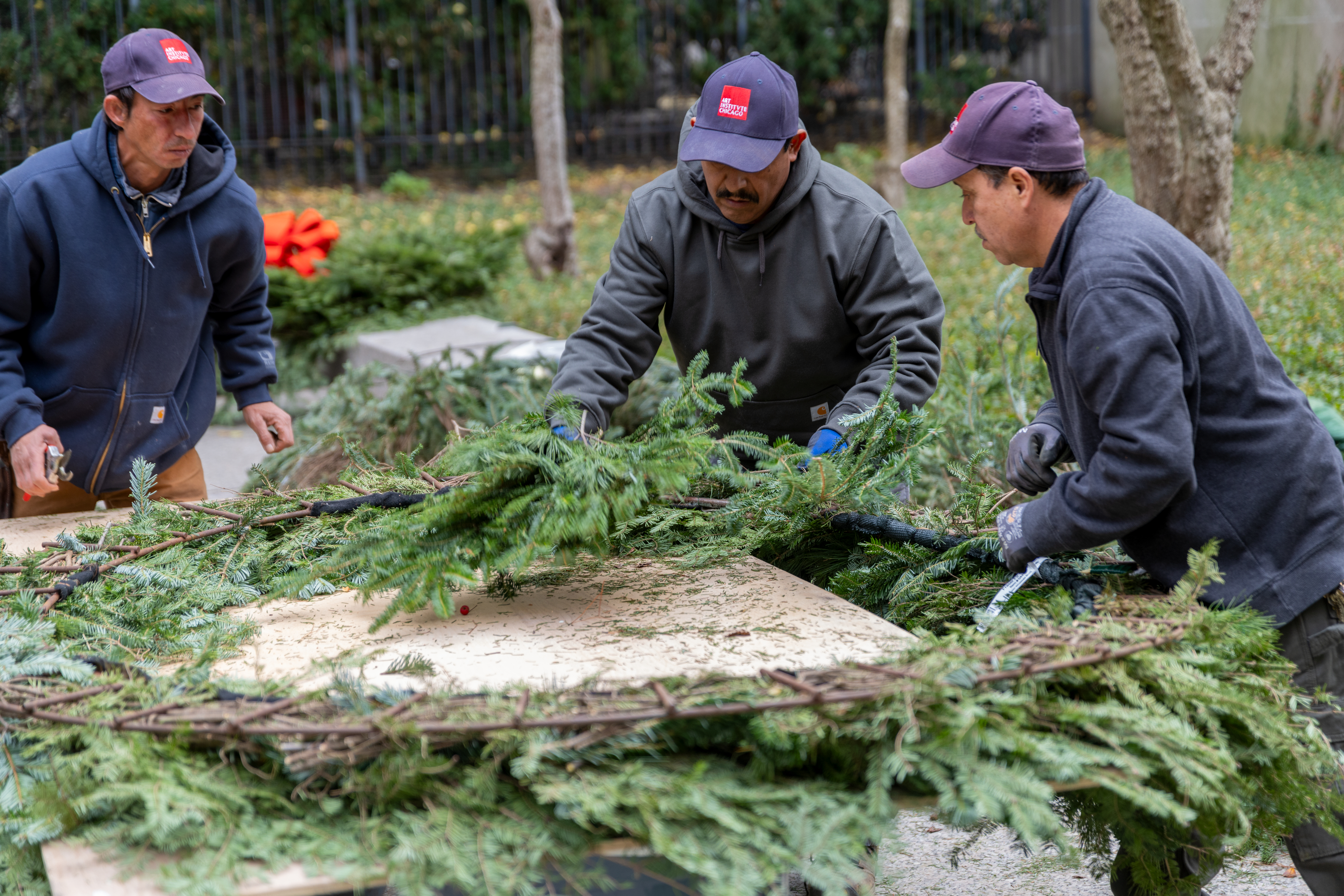 Photo of three men work at an iron form, attaching evergreen branches.