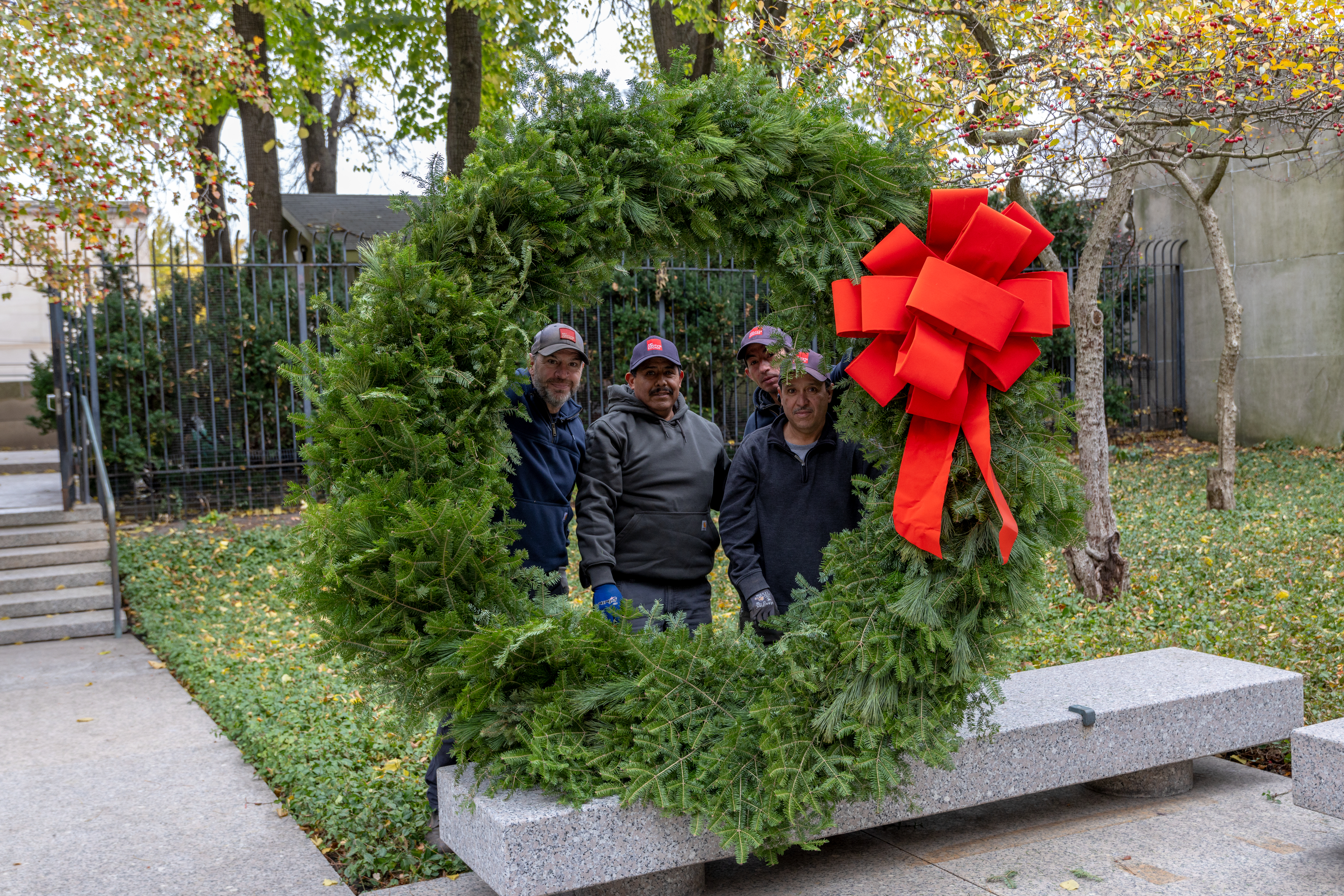 Photo shows four men peeking out from the center of an enormous evergreen wreath and smiling.