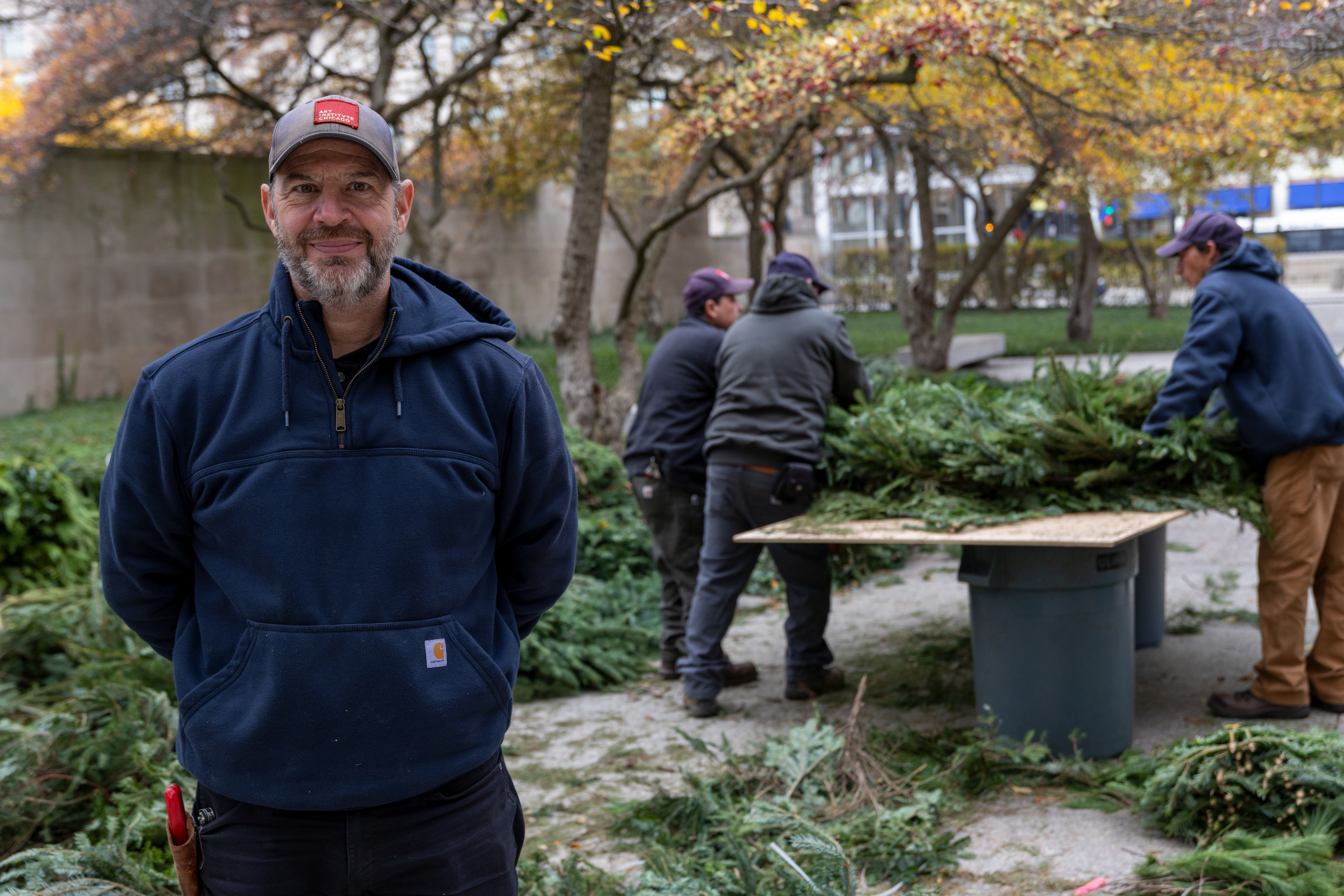 Photo shows a light-skinned man with moustache and short beard, Patrick Thomas, standing outdoors in a blue hoodie facing the viewer. Behind him, three men construct a large evergreen wreath set on a makeshift tabletop.