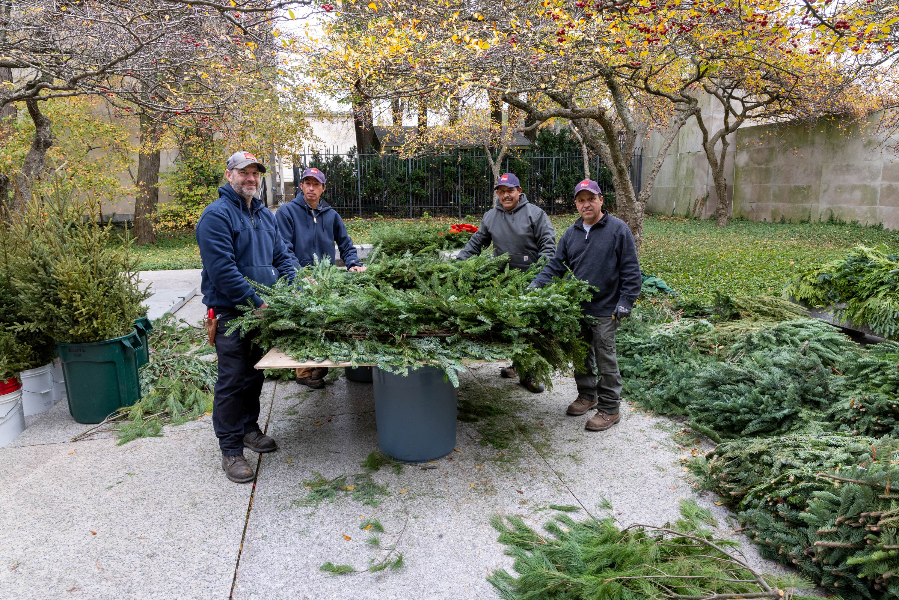 Photo shows four men smiling as they stand around a large, full evergreen wreath set upon a makeshift tabletop in an outdoor garden.