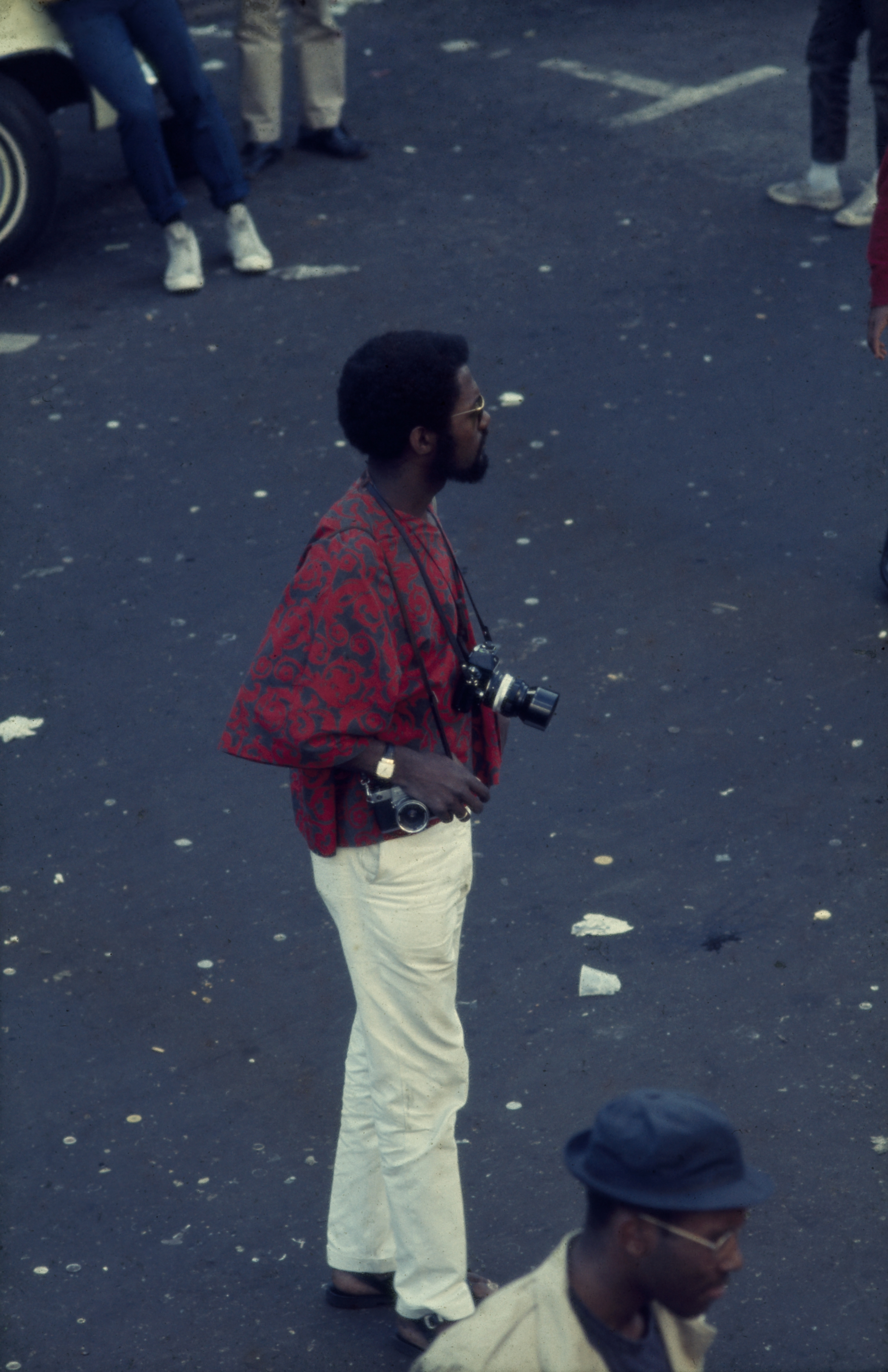 Photograph of a man standing on a paved street, viewed from a slight overhead angle. He wears a red patterned top with wide sleeves, white pants, and sandals. Two cameras hang from straps around his neck, and he holds another camera in his right hand. He has a short afro, a beard, and wears glasses and a wristwatch. Other people are partially visible in the background, including a man in a dark hat at the bottom right. The ground is littered with small debris.