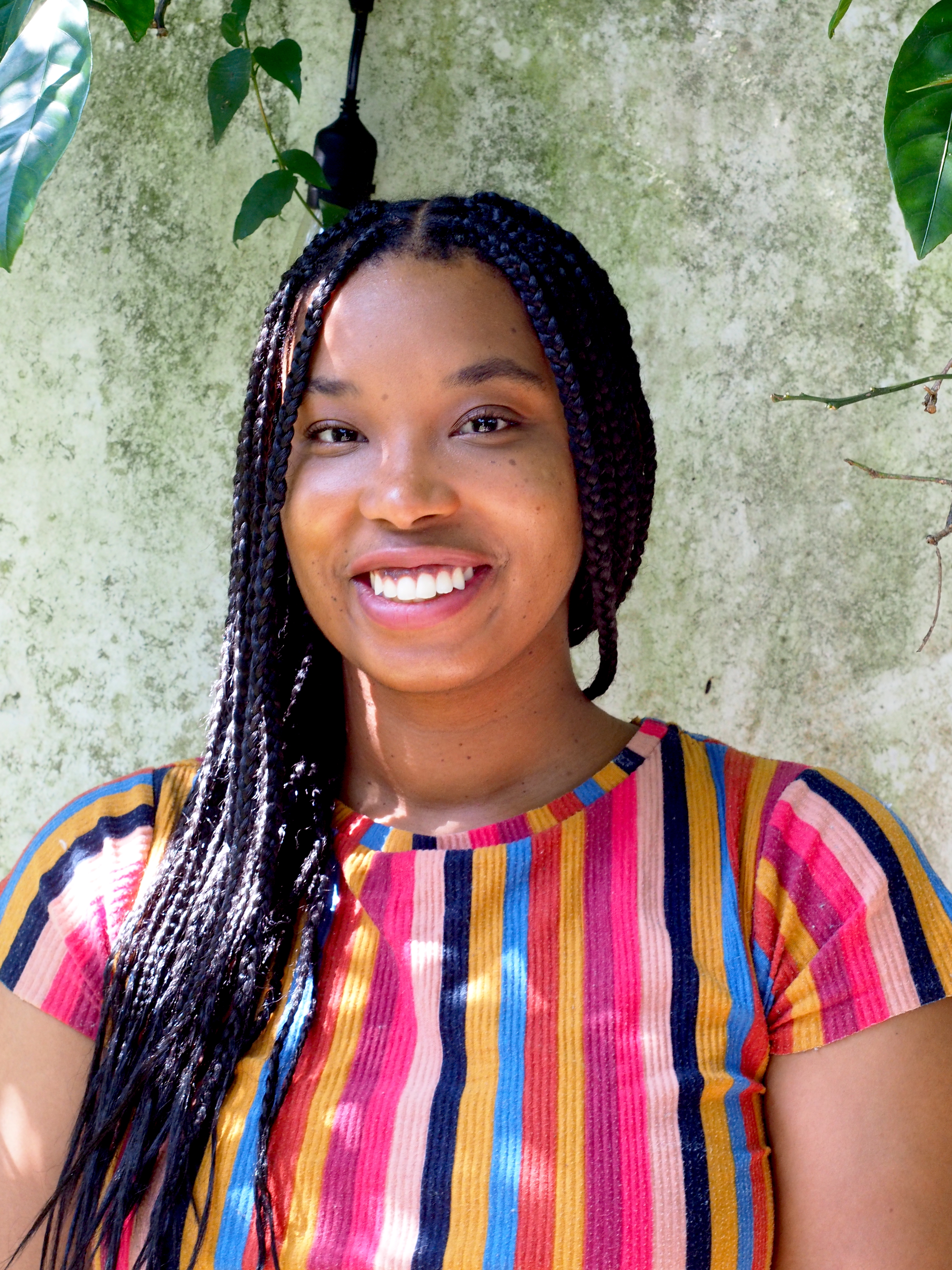 Color portrait of a woman smiling in front of a textured, pale green wall with leafy vines hanging overhead. She has long black hair styled in braids and wears a short-sleeved, ribbed shirt with vertical stripes in pink, red, blue, yellow, and orange. Sunlight filters through the leaves, casting soft dappled shadows across her shirt and shoulder. She faces the camera directly with a relaxed, open expression.