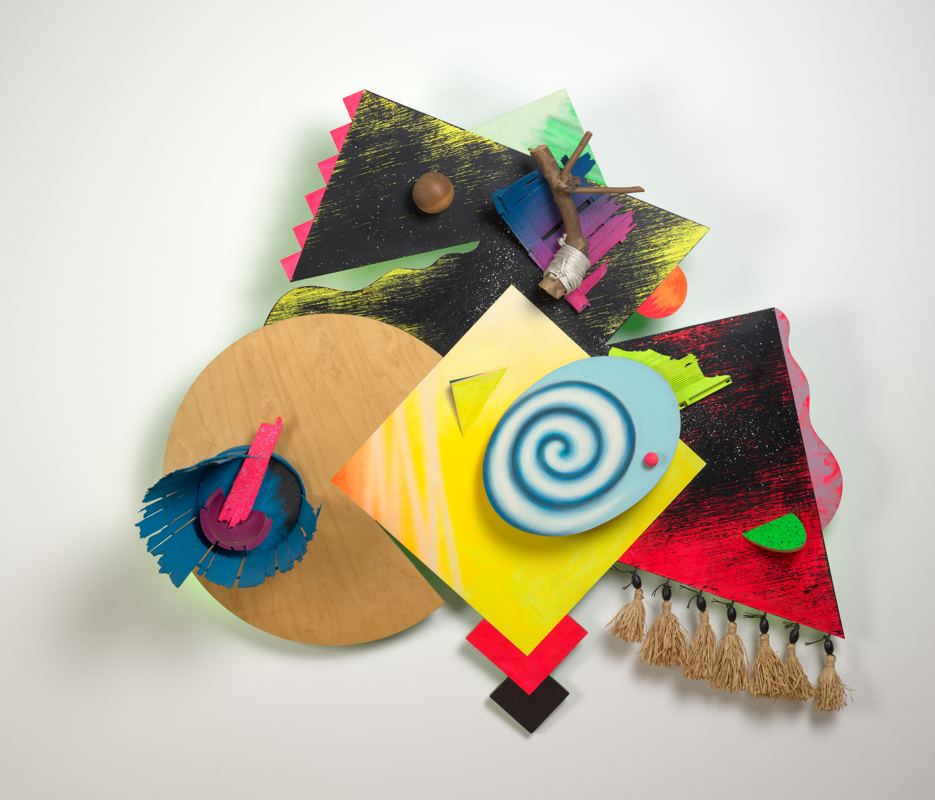 An assemblage composed largely of wooden shapes. At top and center is a rounded, flat shape with a vortex pattern in blue. Below it are numerous other flat shapes that overlap in red, yellow, and black, the woodgrain often visible or enhanced by paint. One shape at right has tassels. A natural-wood circle at left has a blue fringe affixed to it.