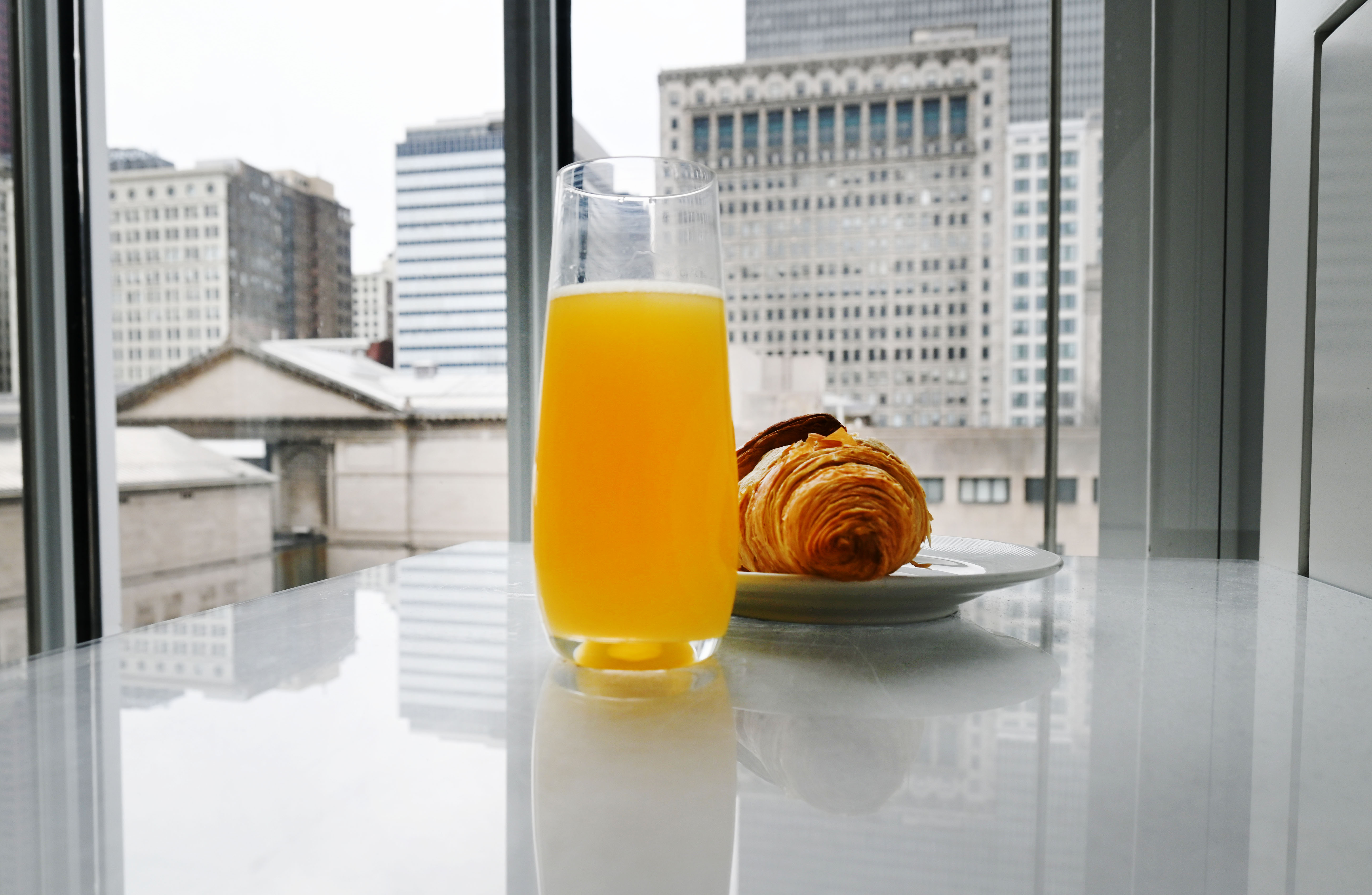 Photo of a tall glass of orange liquid resting on a tabletop before a city skyline, a croissant at right.