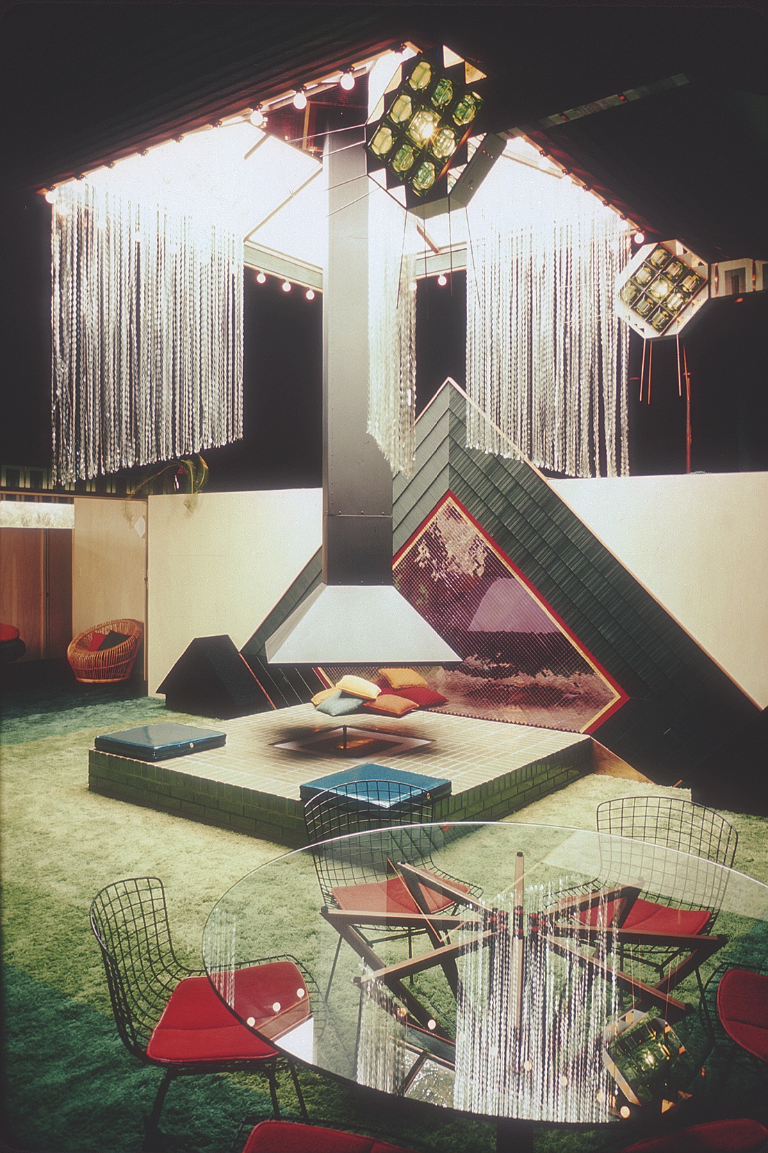 A color photo shows an interior space. The focus is on a raised platform below a large central skylight from which a chimney is suspended. Shimmering strips hang from the double-height ceiling, and a large decorative triangular design is installed on the wall by the platform. In the foreground is a circular glass table surrounded by wire chairs with red seat cushions.