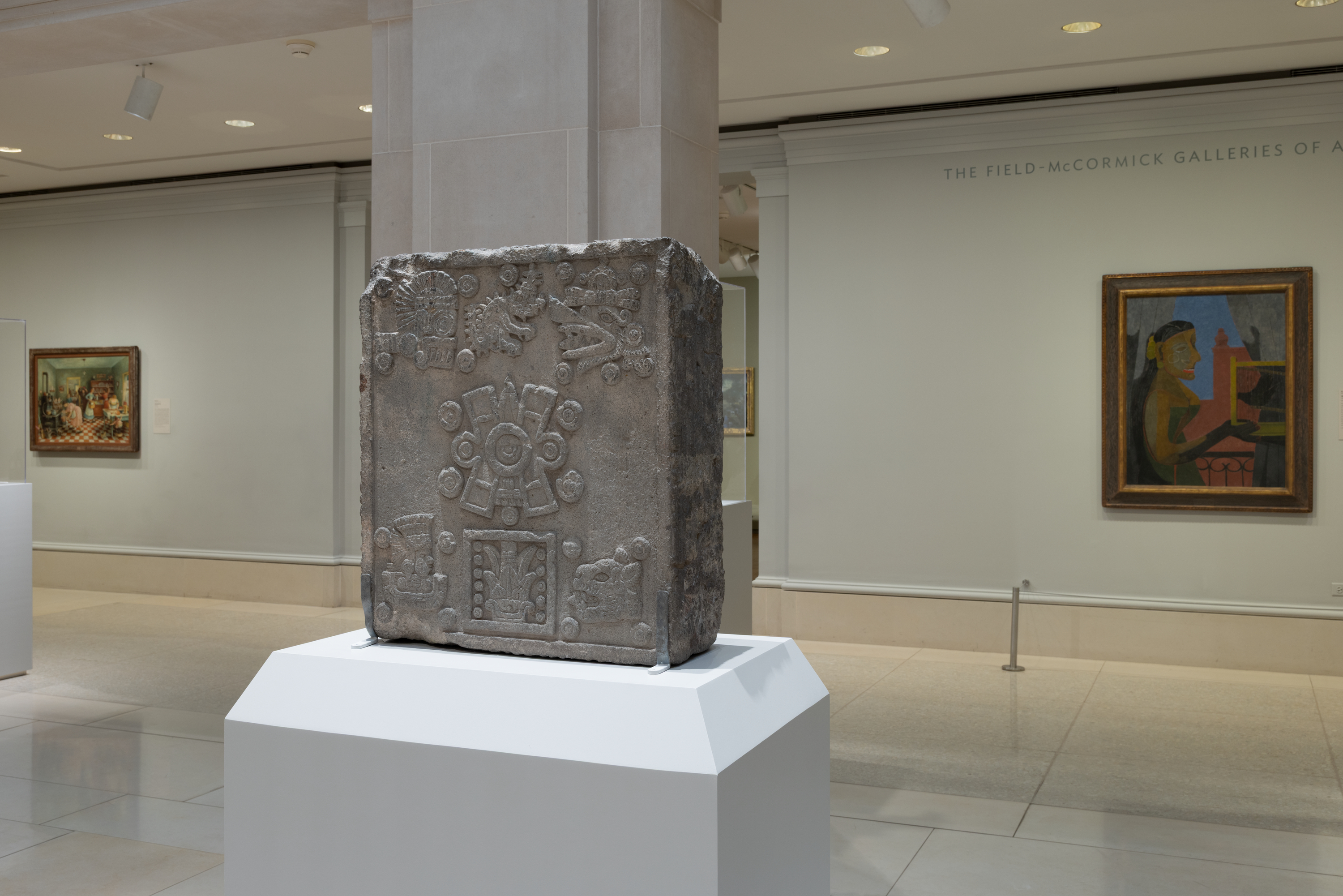 Color photograph of a museum gallery: at center, a gray carved stone block with circles and geometric shapes sits on a white pedestal wrapped around a square column; at left and right, framed paintings hang on pale walls under ceiling lights above a glossy floor.