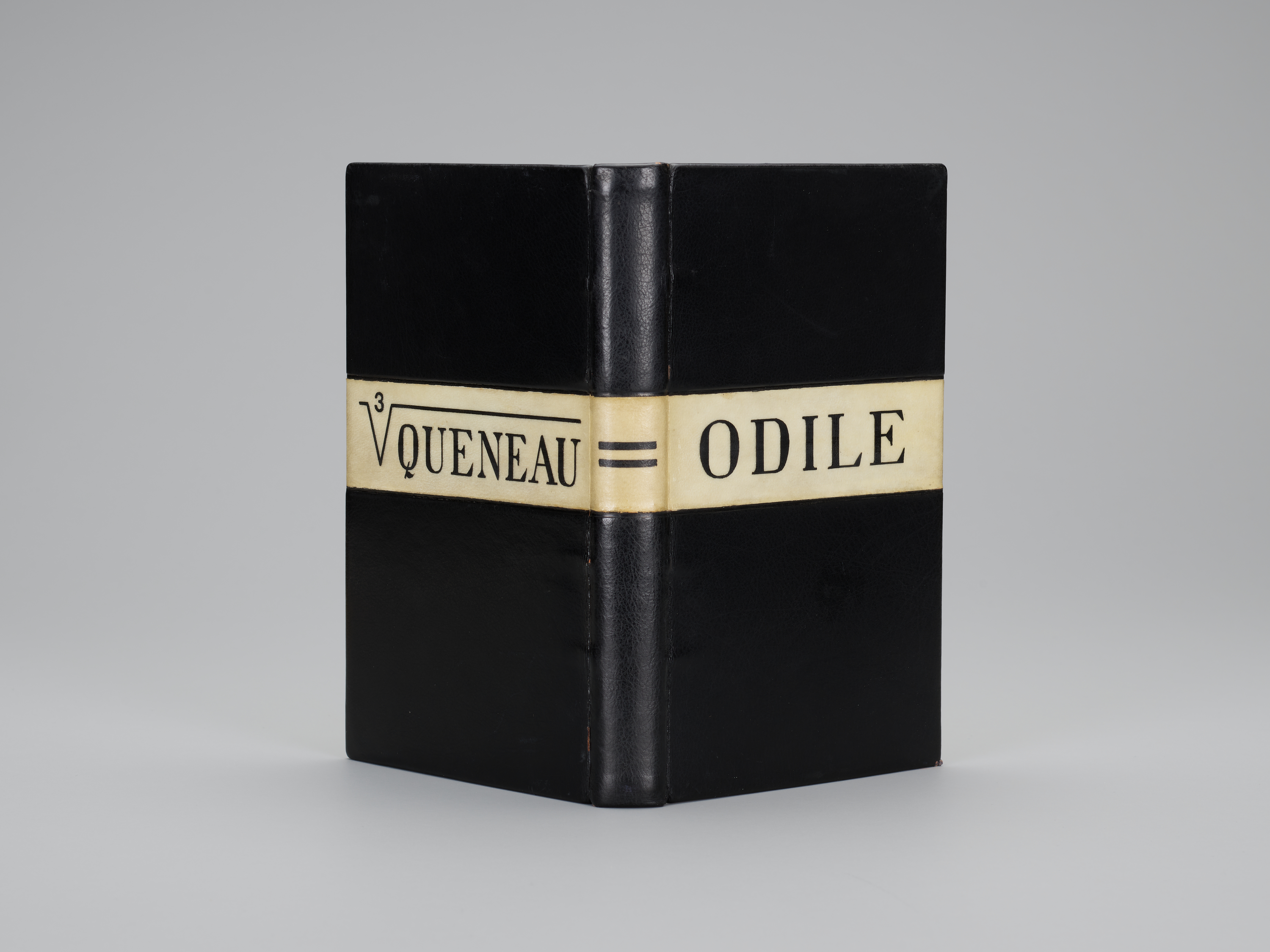 A book propped upright and open, its spine facing the viewer, bound in black leather. A cream-colored band spans both covers and the spine at middle, with a cube root symbol on the back cover over the word "QUENEAU," an equal sign on the spine, and "ODILE" on the front, all in black lettering.