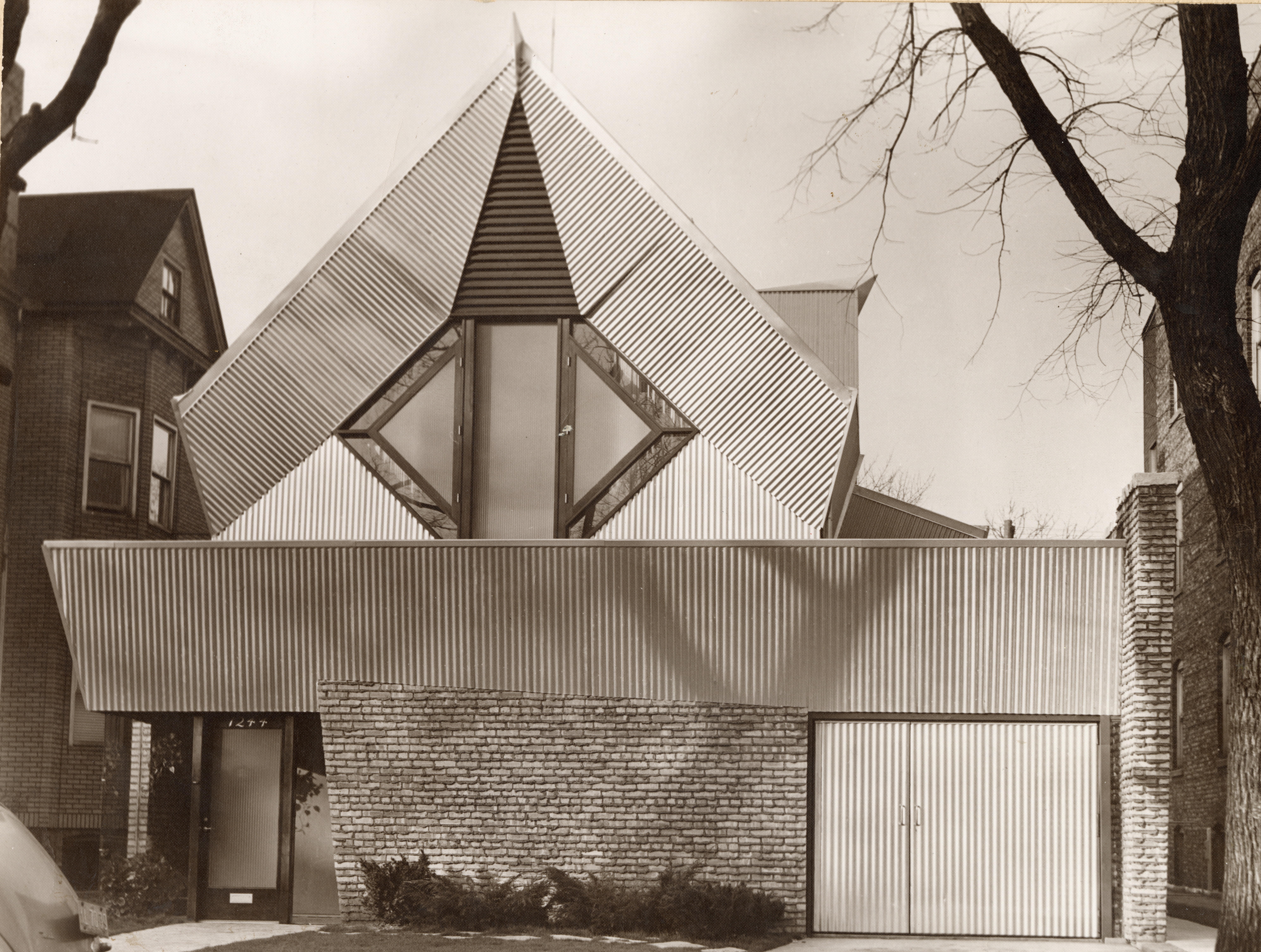 A black-and-white photo of a building. The front single story, with an entrance door and garage, is clad in brick with metal paneling across the top third. A diamond-shape window surrounded by the same metal paneling rises as the building's second story.