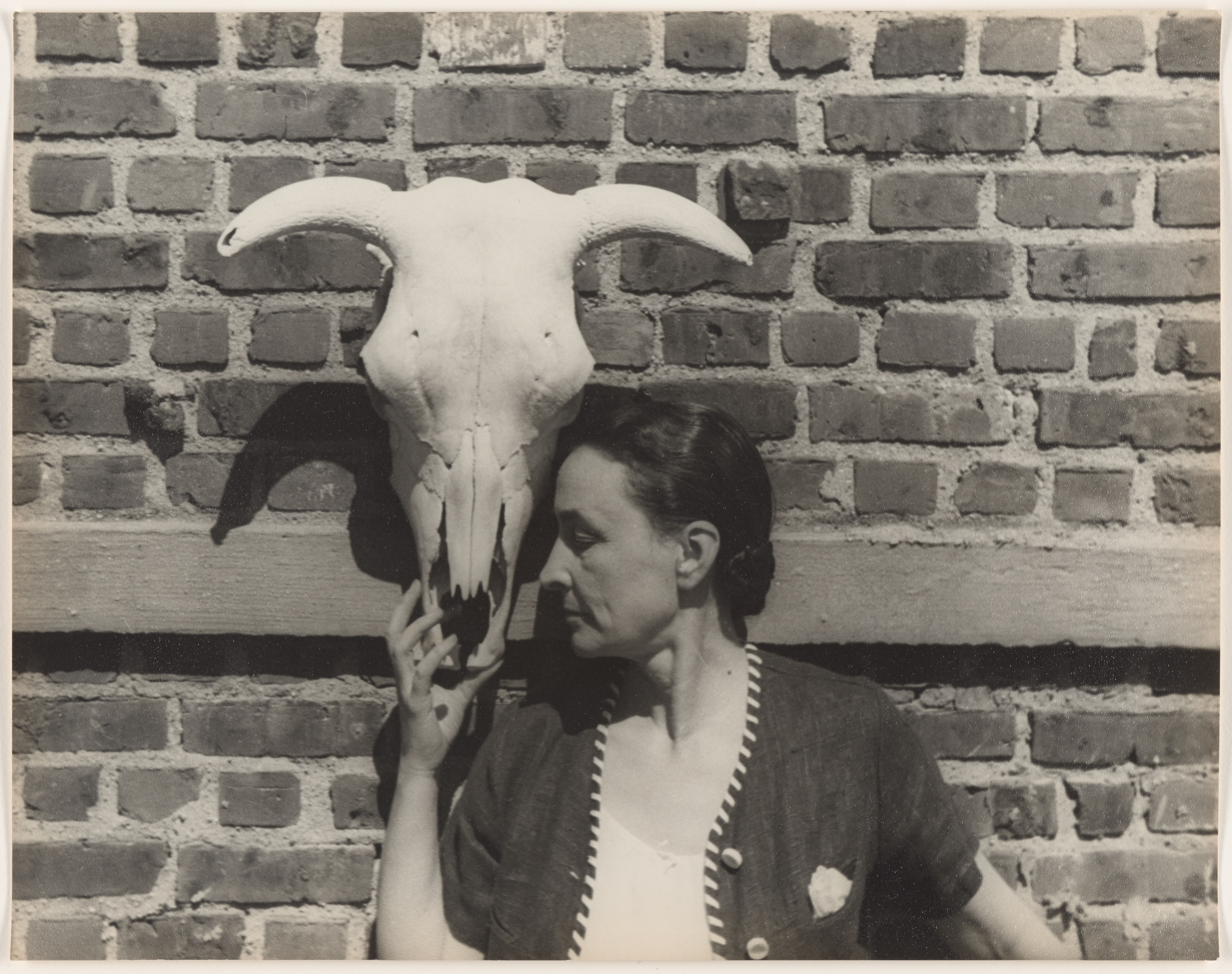 Black and white photograph of a cow skull hanging on a brick wall. A woman leans against the wall, holding its snout in her right hand, her nose pressed against its side.