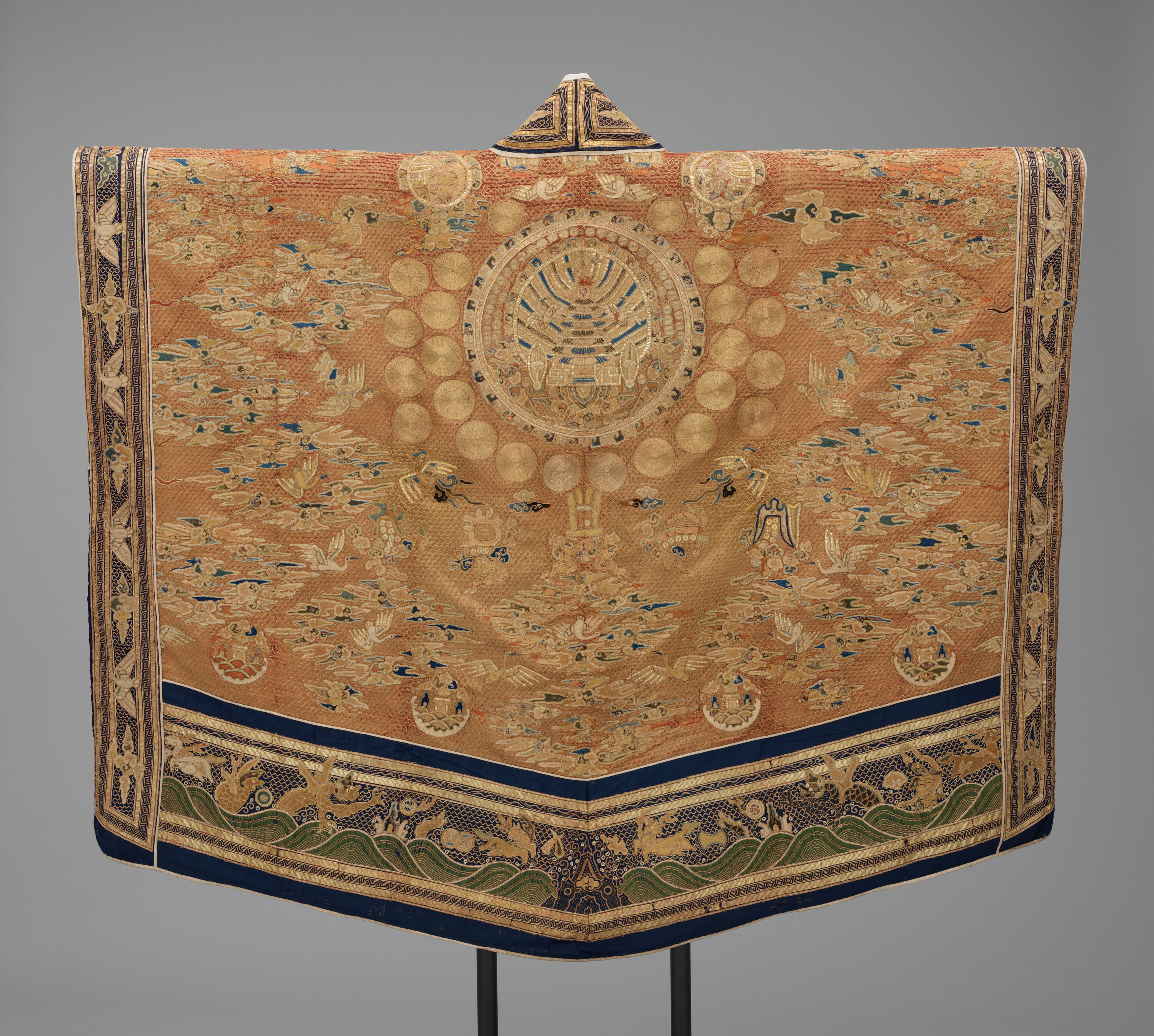 A long, golden-colored robe, held out on a display rod. On it are embroidered tiny birds, medallions, and other embellishments in gold, blue, and black thread.