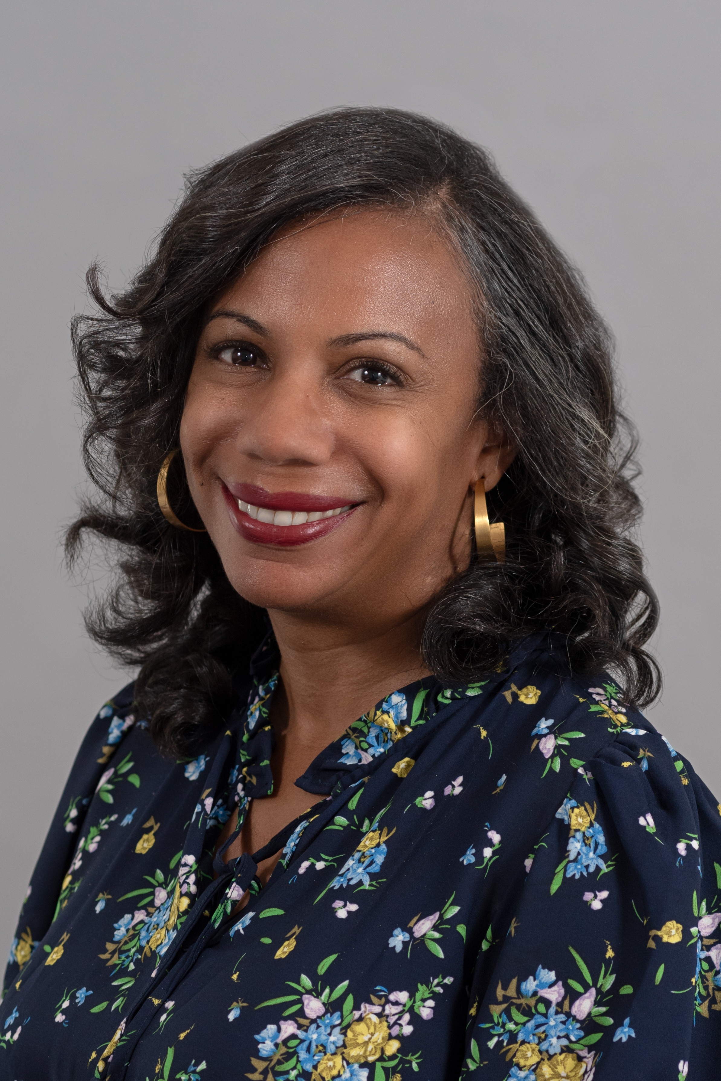 Color portrait of a woman smiling in front of a neutral gray background. She has medium-dark skin, long dark hair styled in soft curls, and wears gold hoop earrings. Her navy blue blouse is patterned with small yellow, blue, and pink flowers. She faces slightly to the left, making eye contact with the camera.