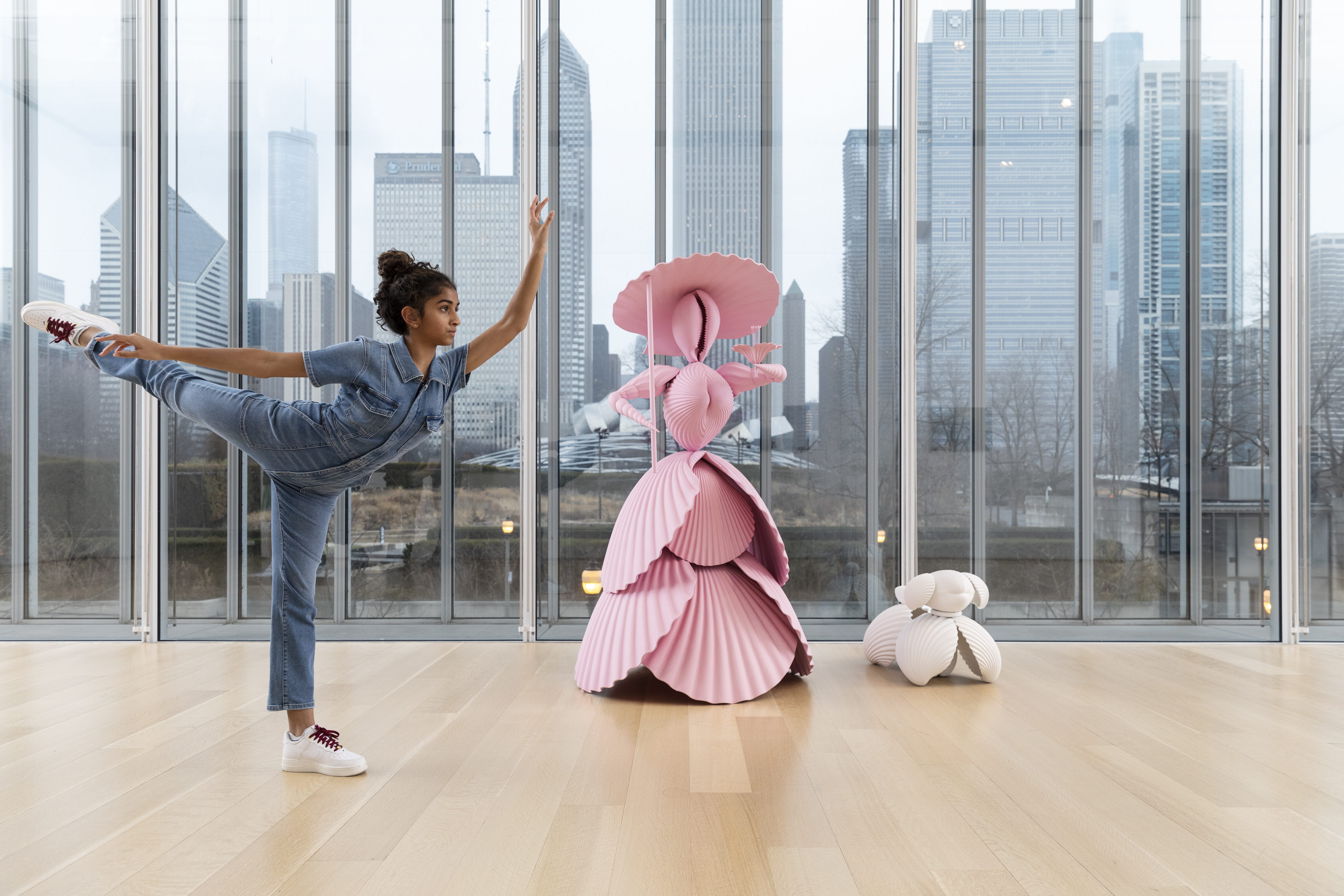 Photograph shows a slim, medium-skinned teenage girl performing an arabesque in an art gallery in front of a life-sized sculpture of a woman and her dog. The women seems to be made of enormous pink shells; her dog, of large white shells. A city skyline can be seen behind the figures.