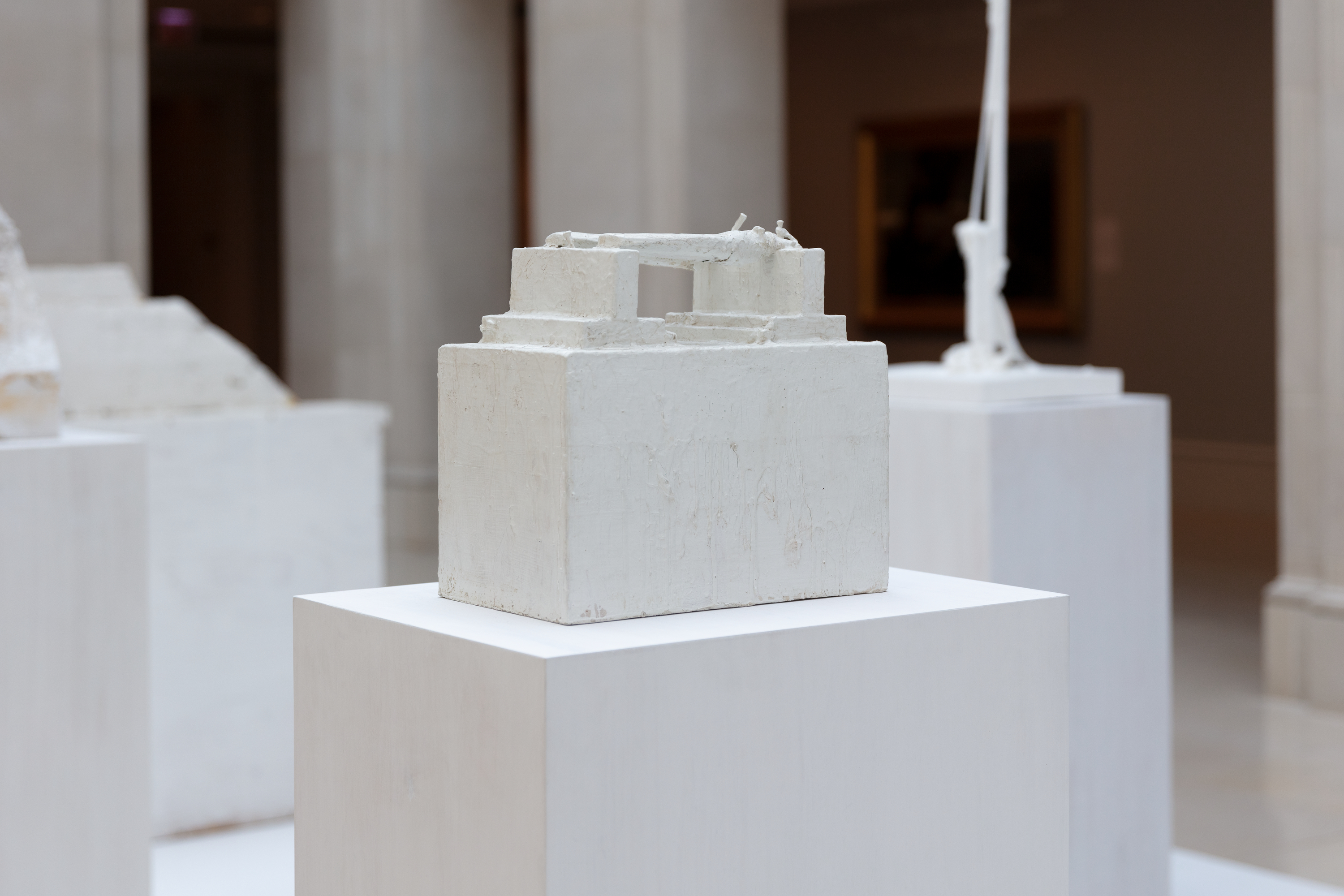 Photo of an art gallery shows a boxy, highly geometric wooden sculpture featuring a wooden box base and stacked, smaller rectangles of wood, the uppermost two spanned by small sticks.