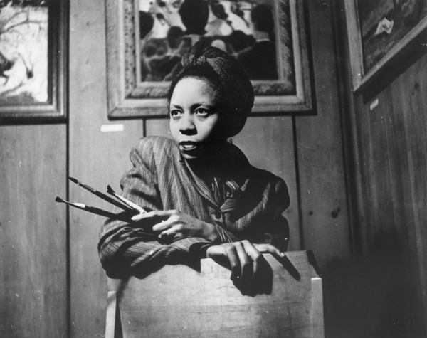 Photograph of a woman seated in front of a wooden wall, holding a set of paintbrushes in her left hand. She wears a pinstriped jacket with a brooch at the collar and has a contemplative expression. Behind her are framed paintings, one of which appears to depict a group of silhouetted heads. The lighting casts dramatic shadows, emphasizing her face and hand resting on the back of the chair.