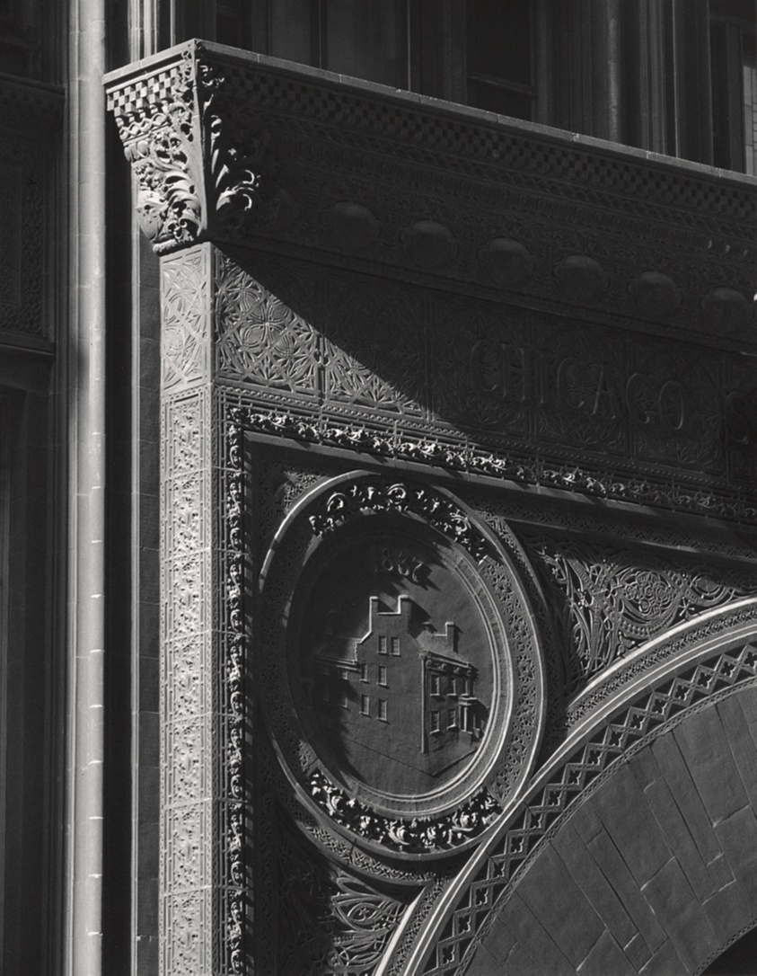 A black-and-white close-up photograph of a decorative architectural element, likely part of a building facade. The image focuses on intricate carvings of floral patterns, with the word "CHICAGO" etched into the stone. Below this is a circular relief depicting a house, with the year "1837" inscribed beneath it. The image emphasizes the ornate details of the stonework, with light and shadow creating a striking contrast.