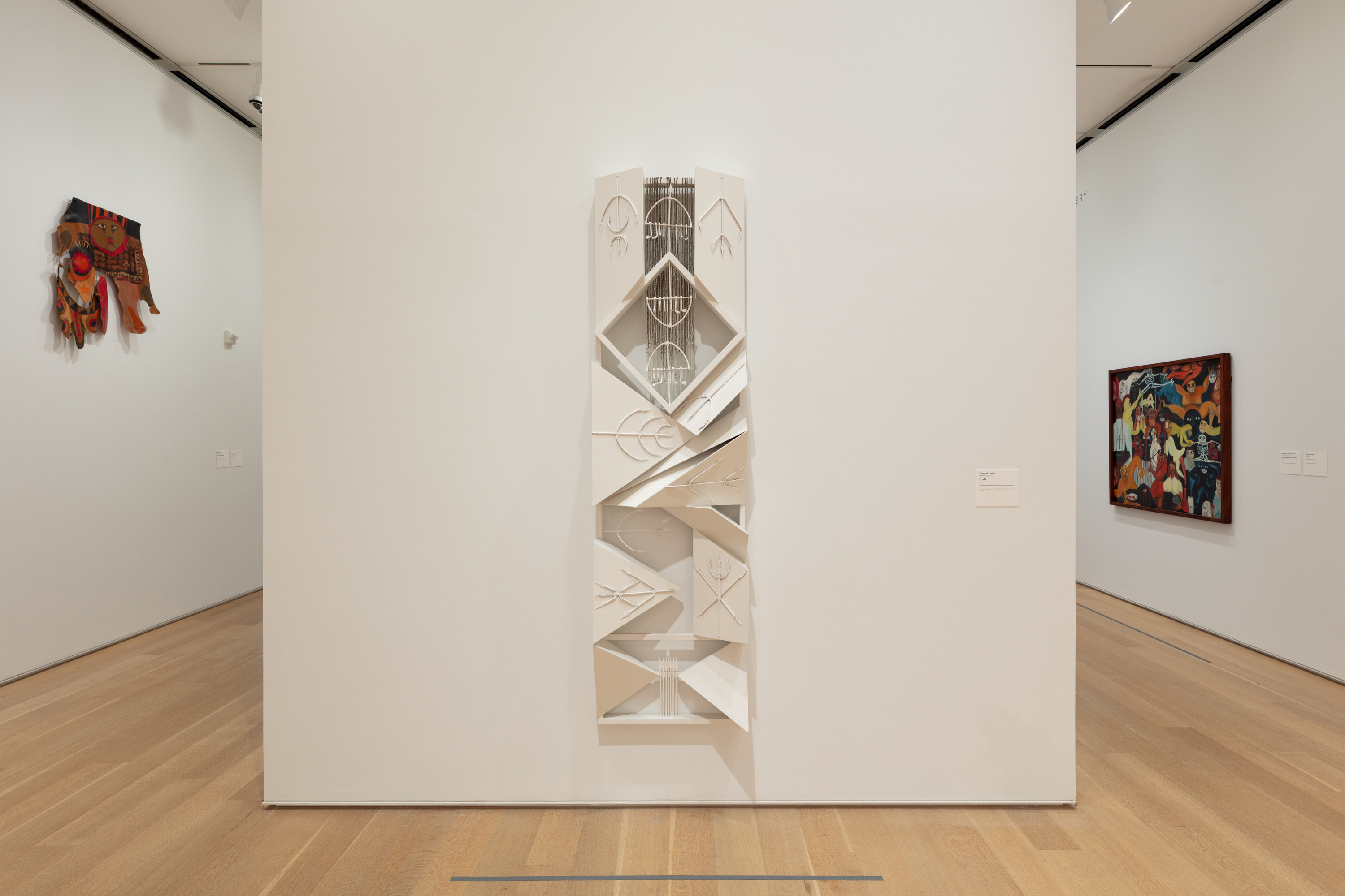 Photo of a white, rectangular sculpture, tall and vertical, affixed to a white gallery wall. The sculpture is composed of many geographic shapes in relief, including a prominent diamond shape toward the top. Accents resembling multipronged spears in some cases are affixed to the surface throughout.