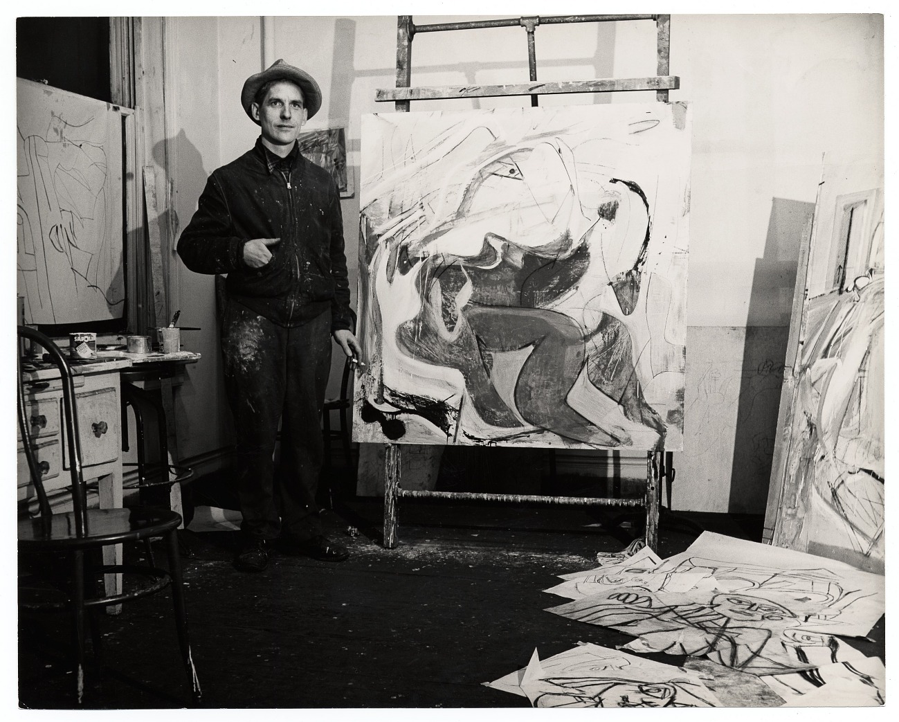 A black-and-white photograph shows a large abstract painting on an easel at center, with an adult wearing a hat and paint-splattered clothes standing at left, a small table with brushes and cans at far left, more canvases at right, and sketch sheets scattered on the floor at bottom.