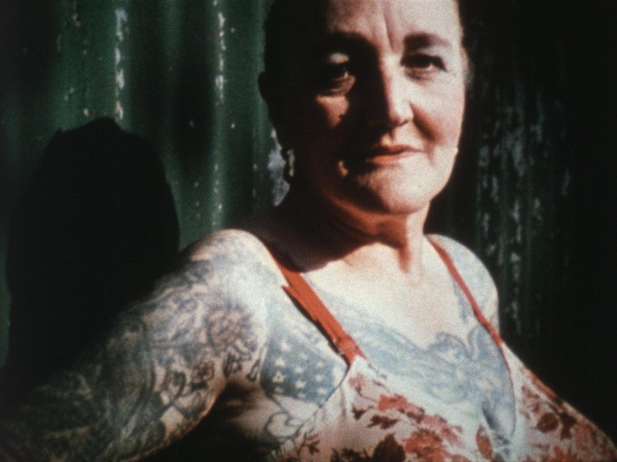 An older white woman with short dark hair stands outdoors in sunlight, facing the camera with a neutral expression. She wears a floral-patterned tank top that reveals detailed tattoos covering her arms, shoulders, and chest, including an American flag motif and floral designs. A green, corrugated metal wall fills the background, casting a deep shadow beside her.