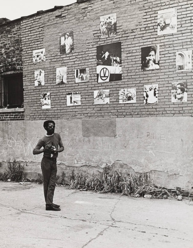 A Black man stands in an empty lot beside a brick wall, holding a camera and looking up at a series of photographs displayed on the wall. The photographs, arranged in an informal grid, include portraits, protest scenes, and community moments. The lower part of the wall is covered in concrete, with faint graffiti visible. Grass and weeds grow along the base of the wall, and the sidewalk is cracked.