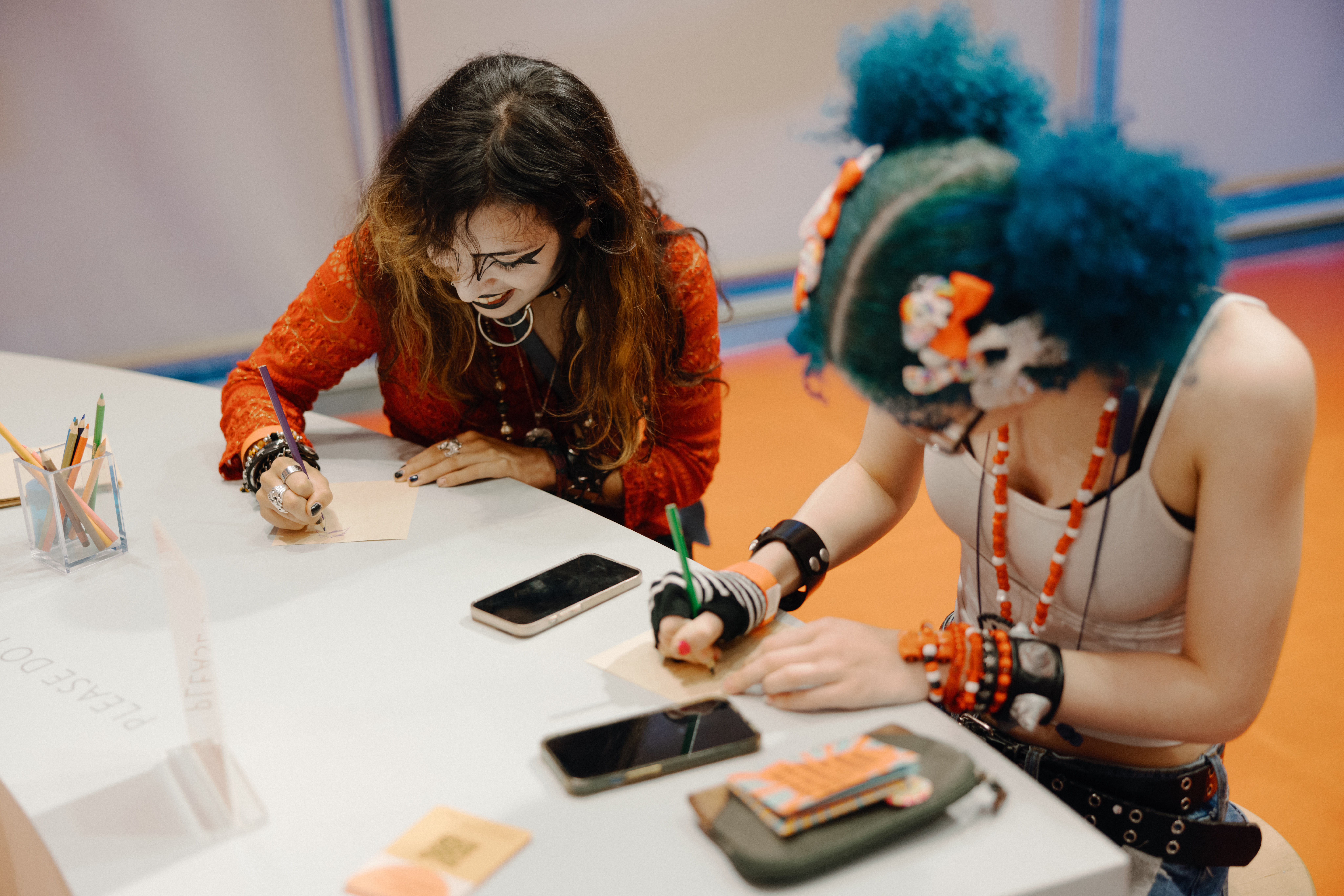 Photo shows two light-skinned teens sitting at a table drawing. One has voluminous blue hair in pigtails adorned with neon-orange bows and white flowers. The other wears black lipstick.