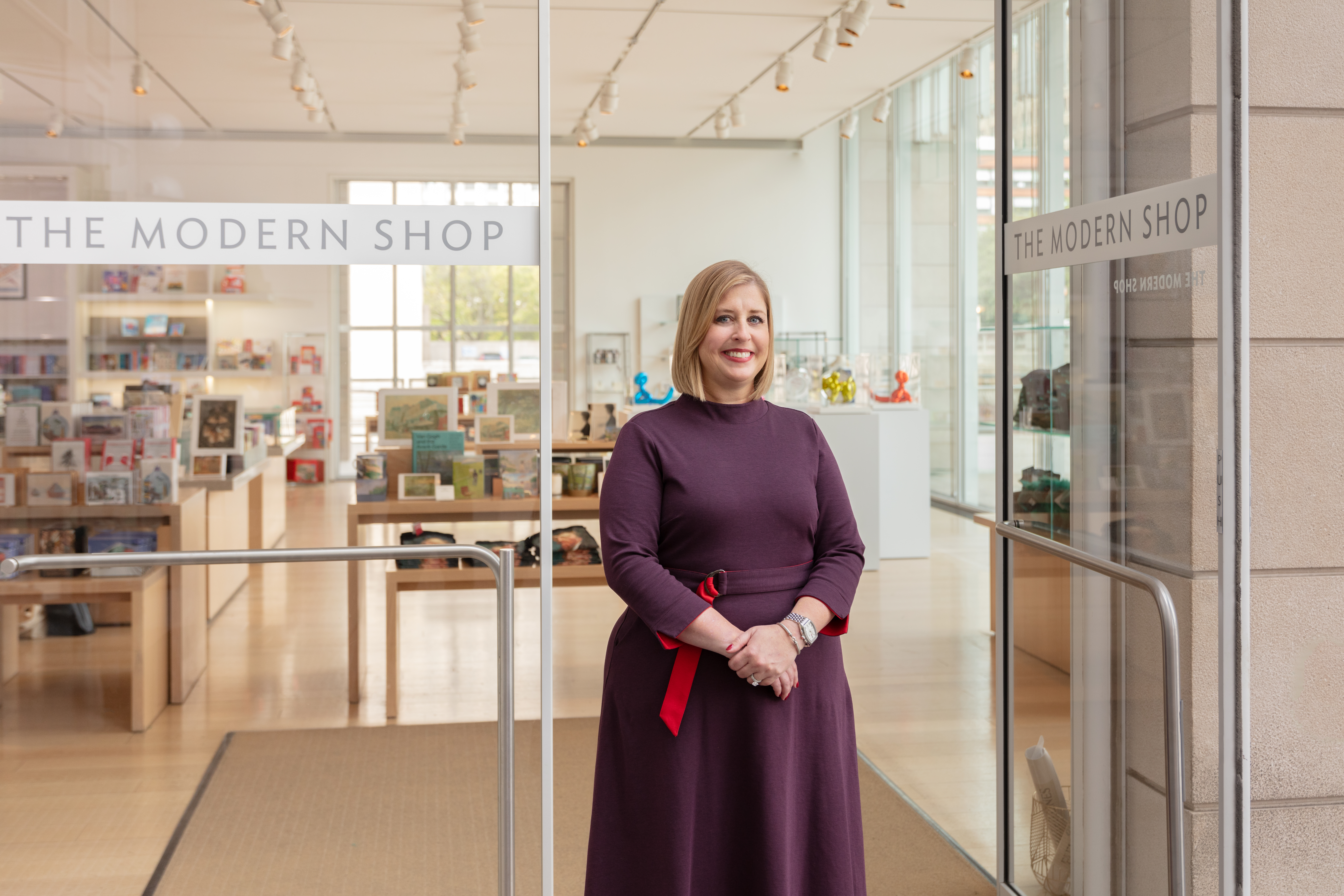 A light-skinned blonde woman, Heather Reinholtz, stands in the entryway of the Art Institute of Chicago's shop in the Modern Wing, the words "The Modern Shop" written in block letters on the glass door to her left. She wears a burgundy dress. Behind her, through the doors, is a sun-drenched gift shop filled with books and other items.
