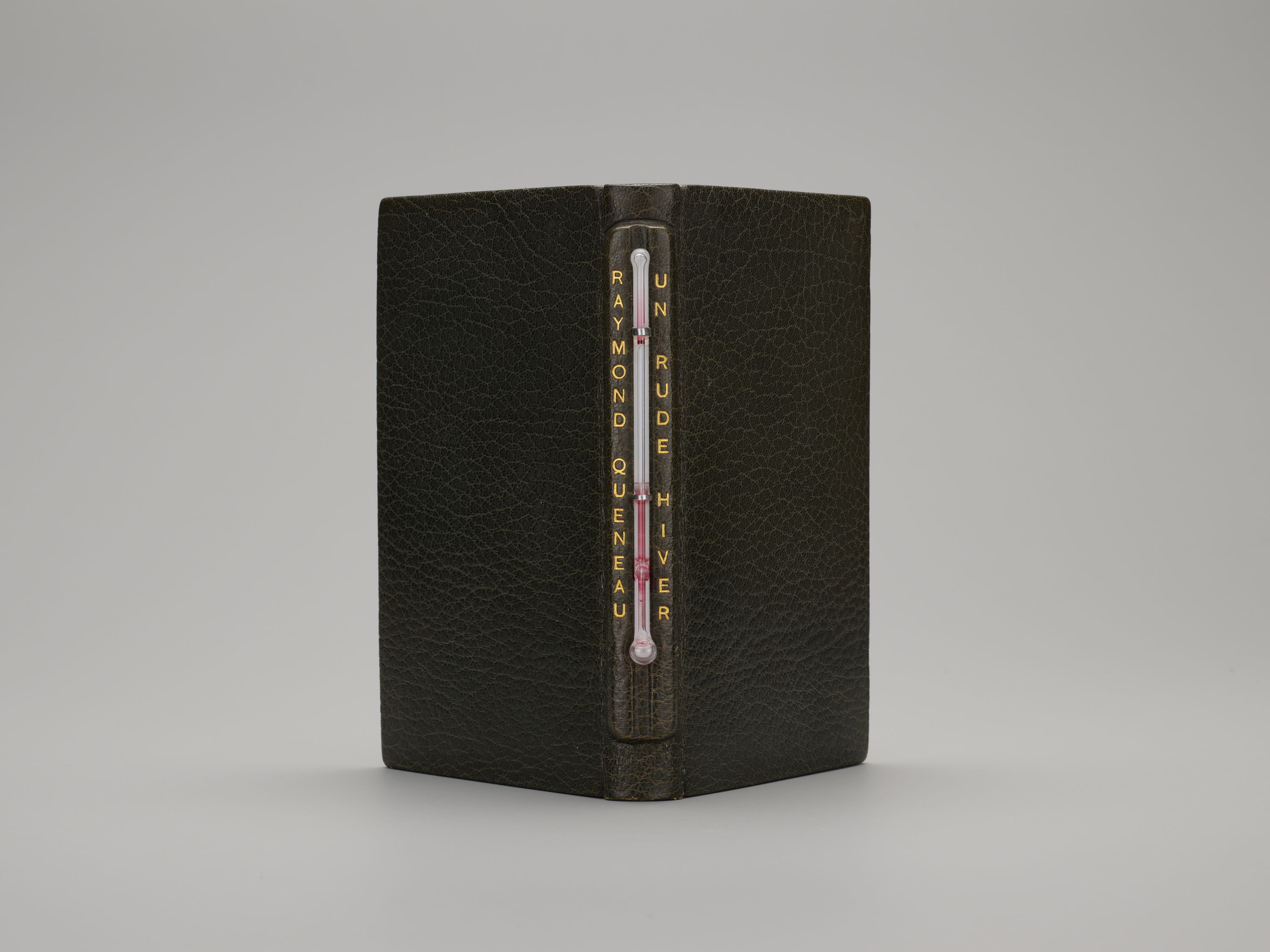 A book propped upright and open, its spine facing the viewer, in black pebbled leather with a mercury-filled thermometer embedded into the spine. Gold lettering on the spine reads "RAYMOND QUENEAU" and "UN RUDE HIVER."