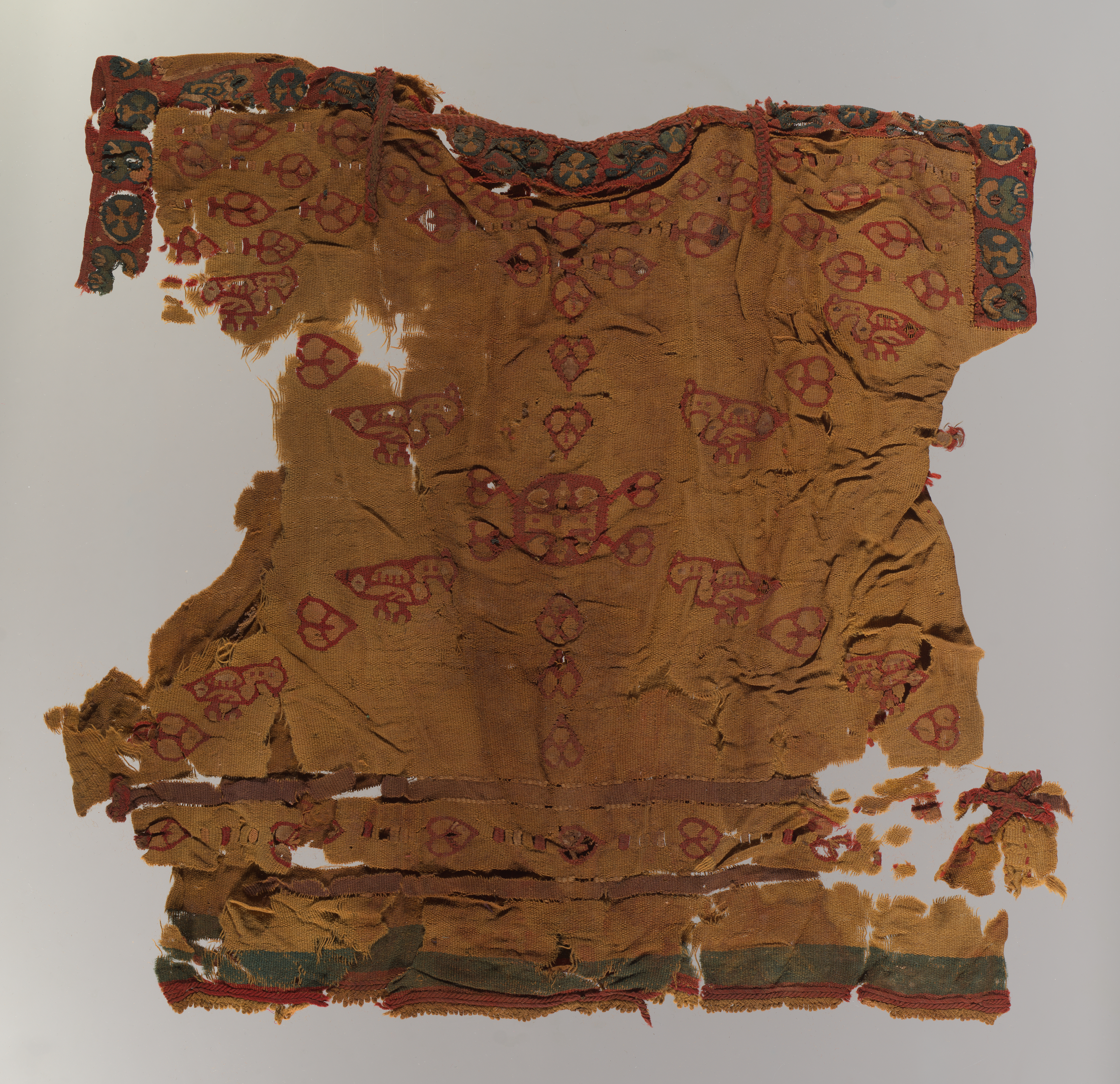 A tattered and heavily degraded brown garment that was once a tunic and still holds some semblance of that shape. It sports a motif of birds and shapes in red thread and has green and red trim. Many parts of the garment have broken off entirely and have been placed near where they would have originally been attached.