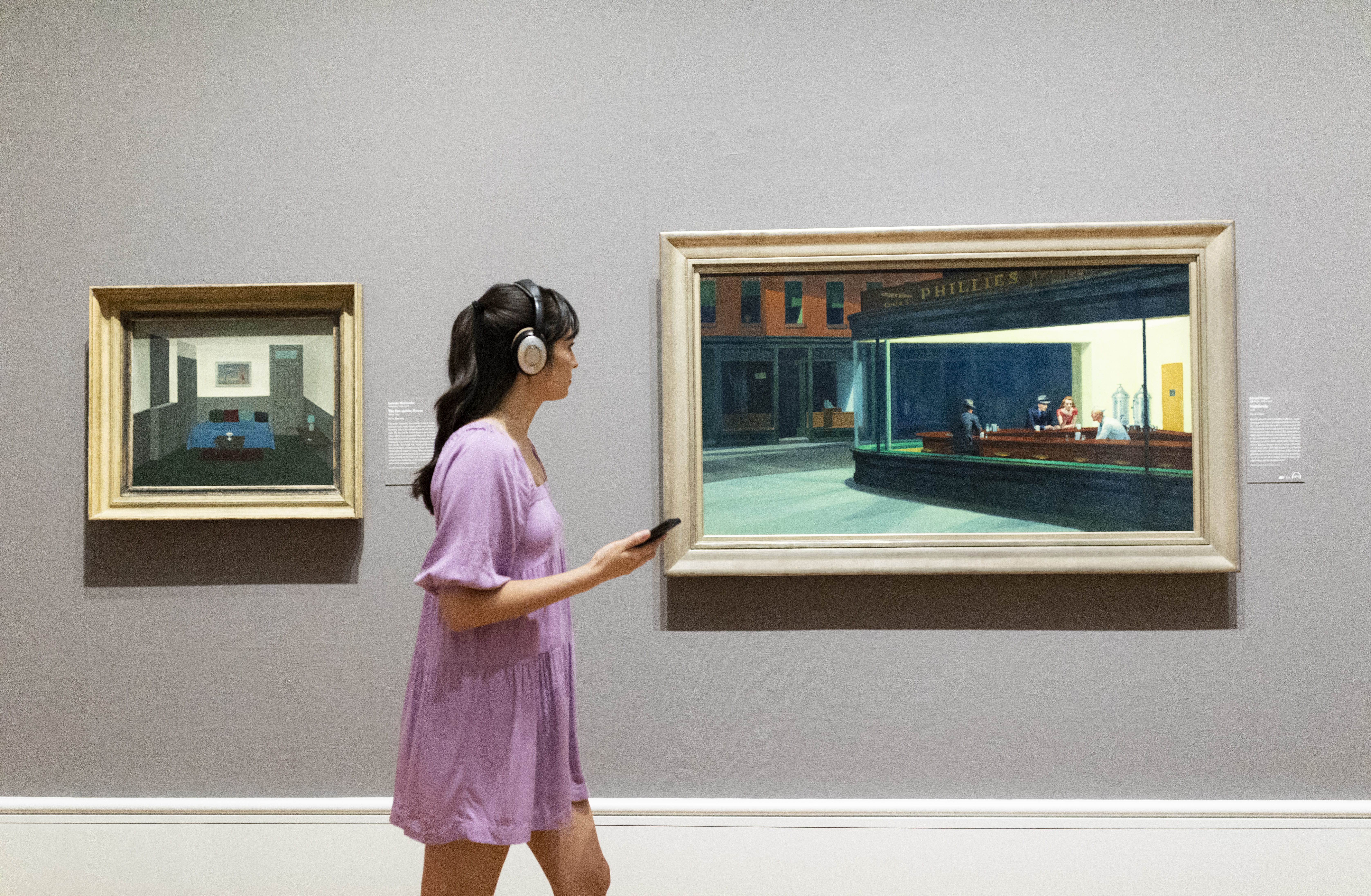 A young woman with light skin and dark hair walks in an art gallery between Gertrude Abercrombie's painting "The Past and the Present" and Edward Hopper's painting "Nighthawks" wearing headphones and a lilac-colored dress.