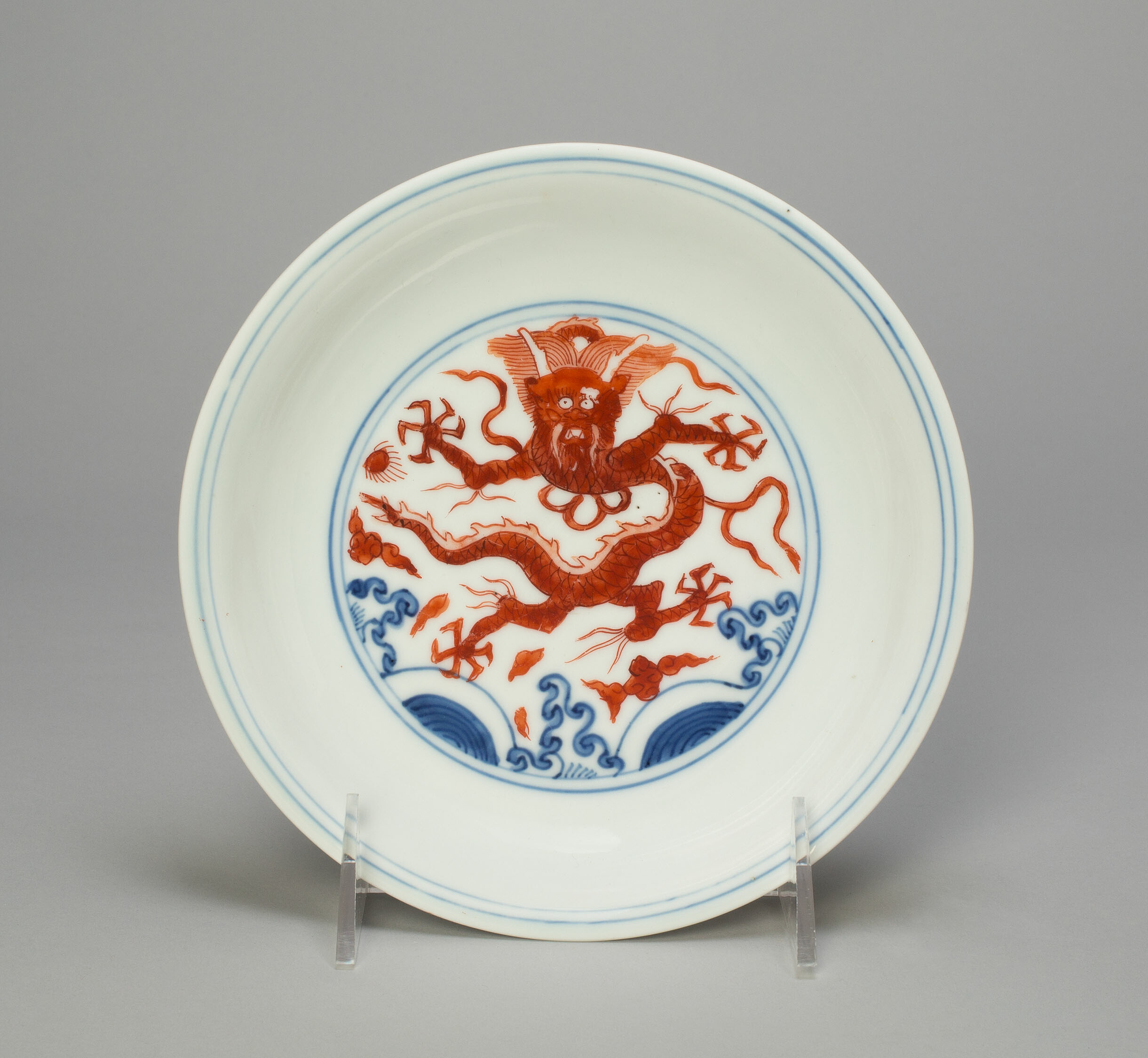 A porcelain plate with light blue edges. From a center circle, a red dragon with pinwheel claws stares out above red clouds and blue waves.