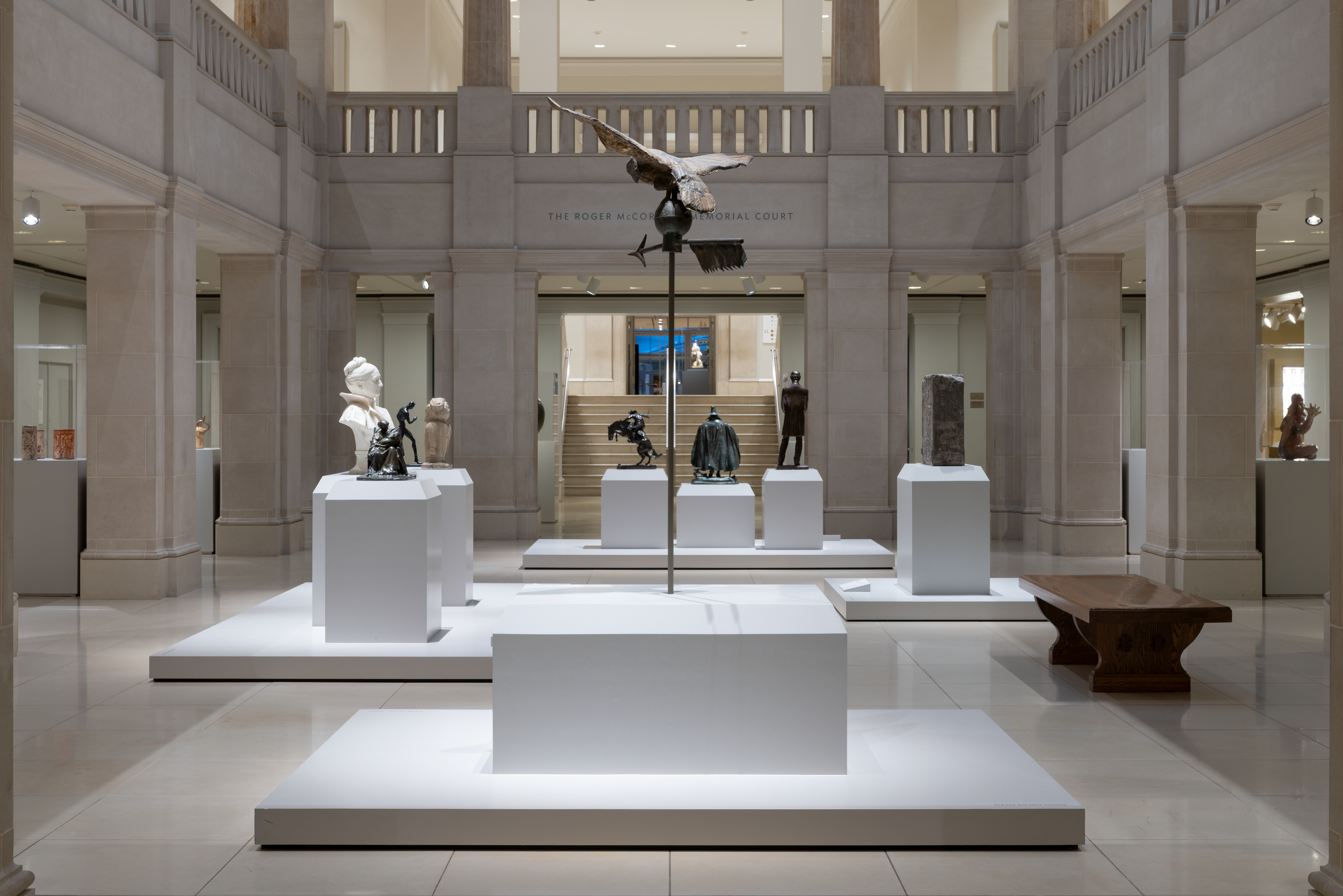 A color photograph of a museum gallery shows white platforms displaying sculptures, dominated by a bird with spread wings mounted on a tall pole at center; at left are a white bust and small figures; at right a wooden bench and a tall rectangular piece; stairs and more pedestals sit in back.