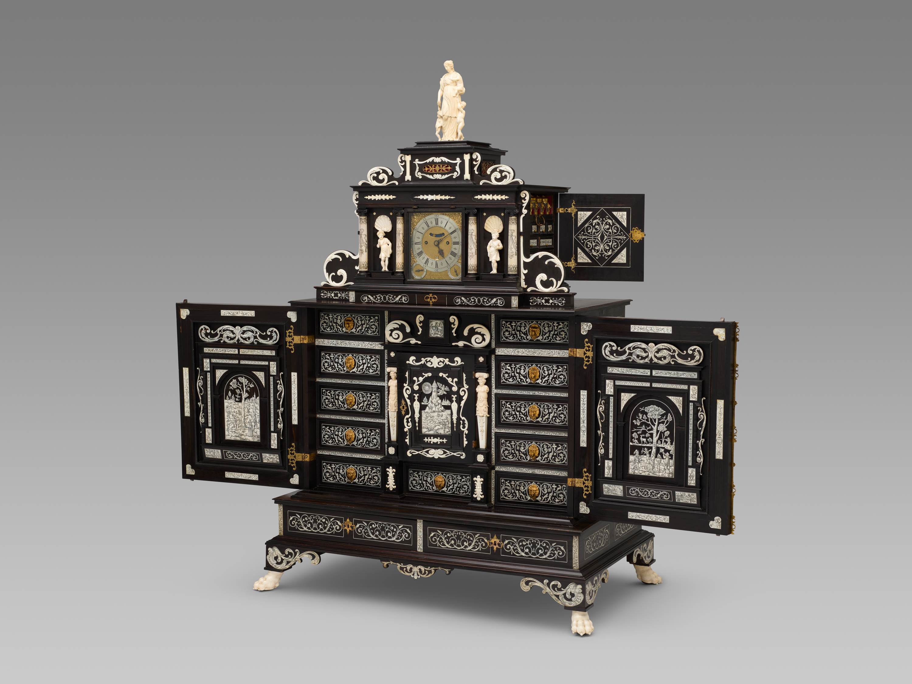 A large black cabinet with intricate white ivory inlaid decorations in scrolling plant-like designs as well as figurative elements. The cabinet is shown in a 3/4 profile with several doors opened to reveal more ivory decoration and drawers inside. The top portion contains a gold and silver clock at its center. Brass hardware serves as both drawer pulls and hinges, and ivory lion paw feet sit at each of the bottom corners.
