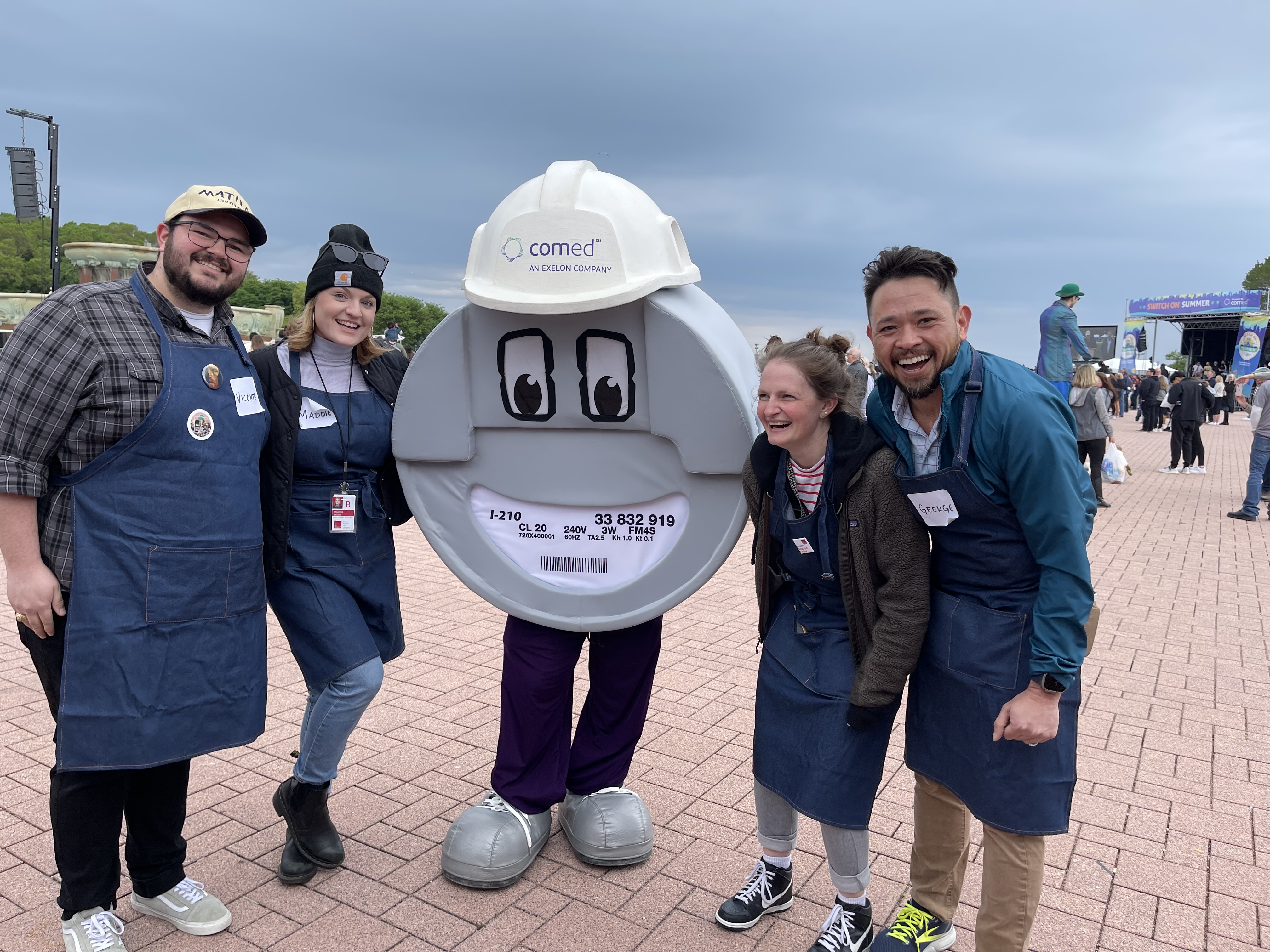 A photo taken outdoors on an overcast day shows 4 people, Maddie Shearer among them, posed and smiling with a figure dressed as a cartoonish version of an electrical meter.