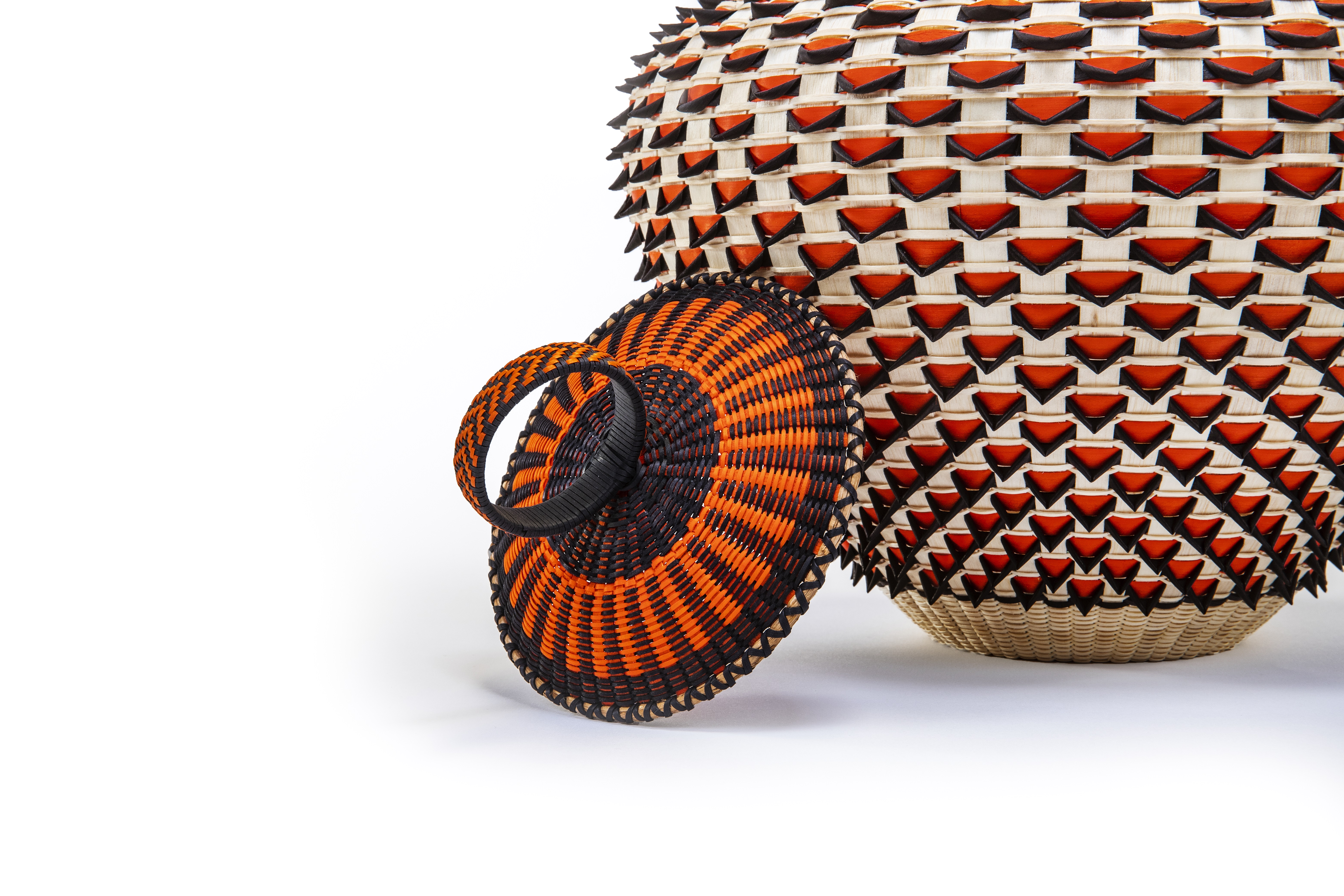 Close-up view of a round basket lid woven in bright-orange and black. Behind it is the partial view of an ash basket made of blond, red, and black wood, the tight weaving pattern making small upside-down triangles out of the red.