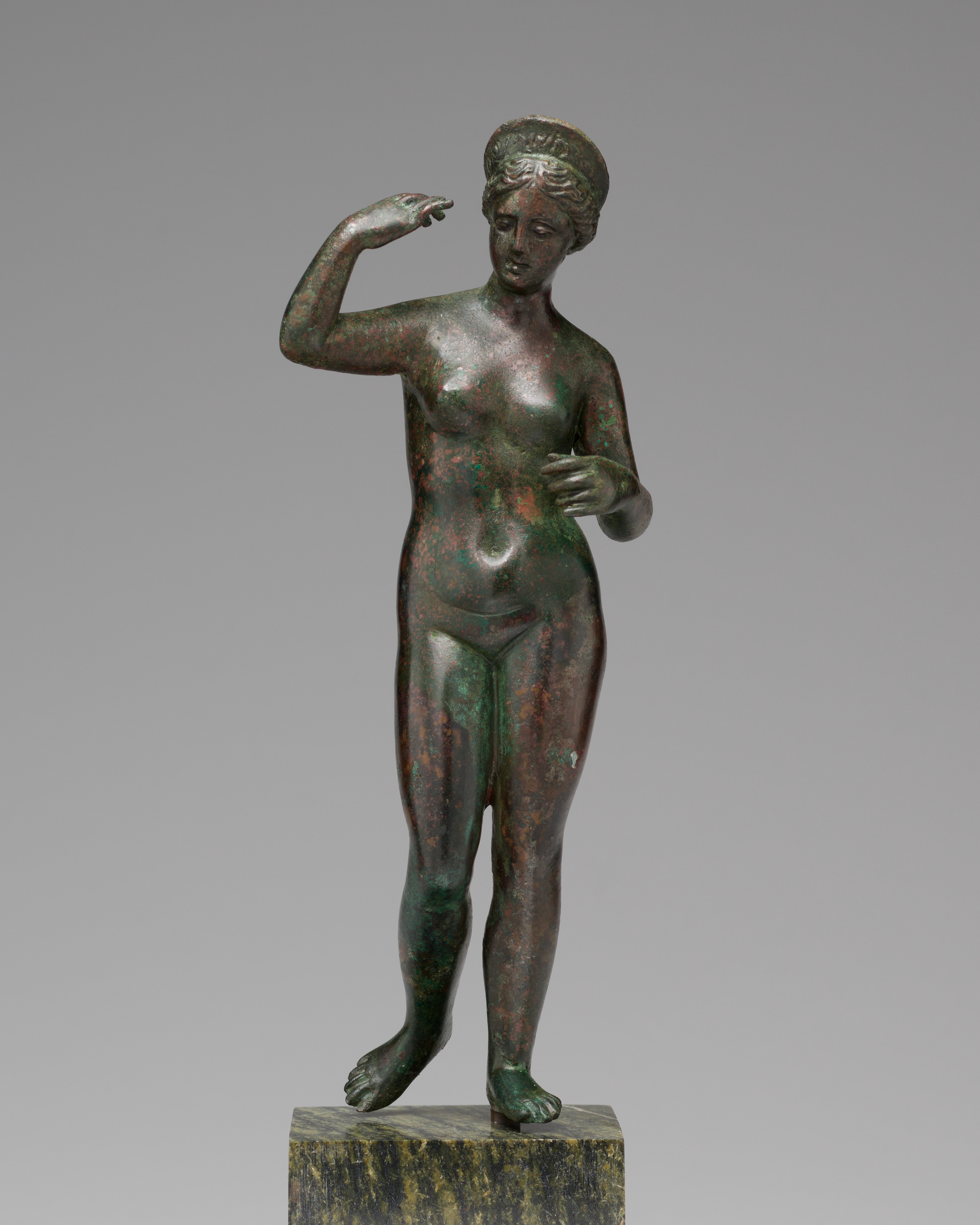 Frontal view of a bronze sculpture of nude woman on narrow pedestal, her hand raised. One foot steps forward. She looks to the ground.