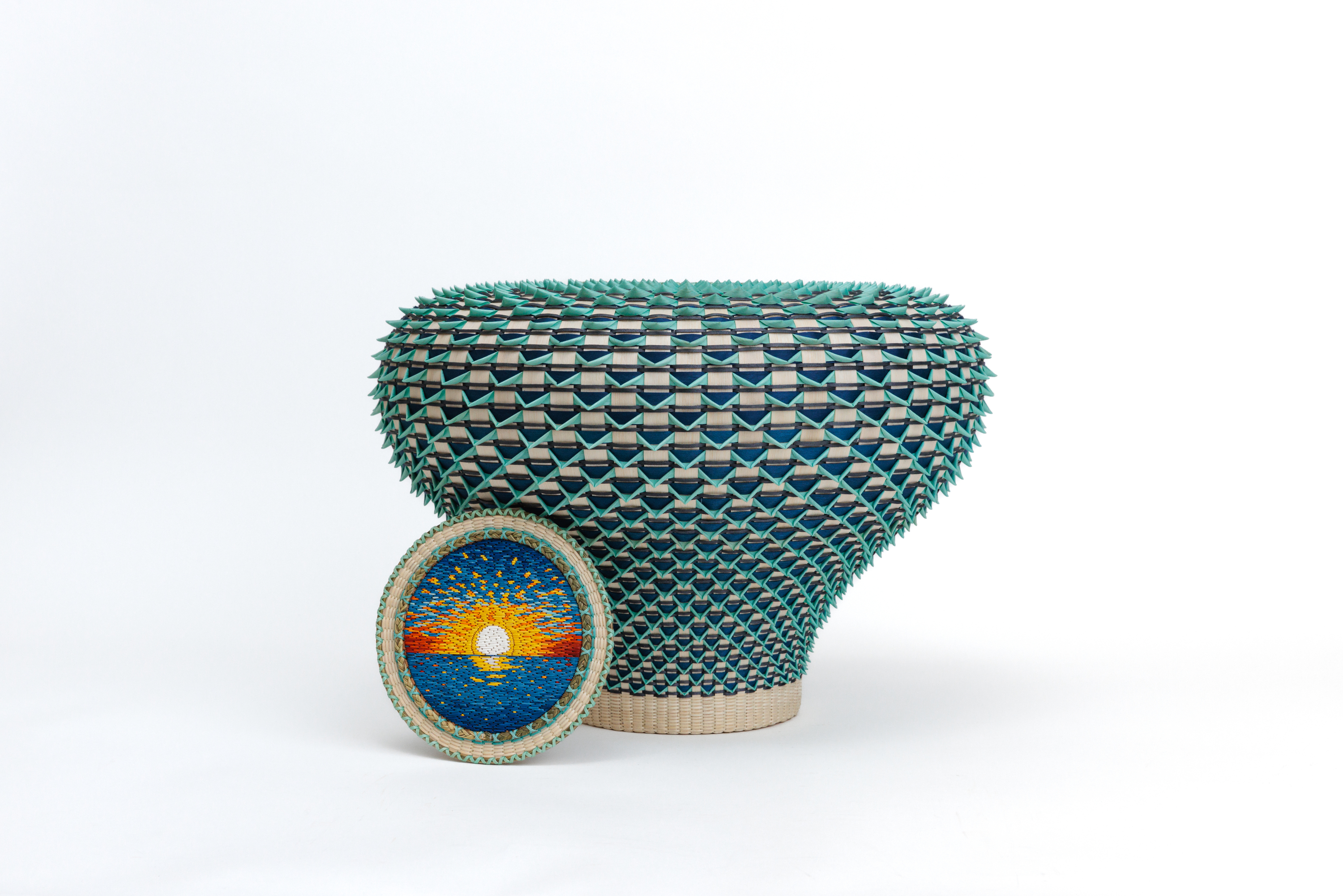A woven basket in blond and blue-green wood with a lid, placed to the side, featuring a woven horizon.