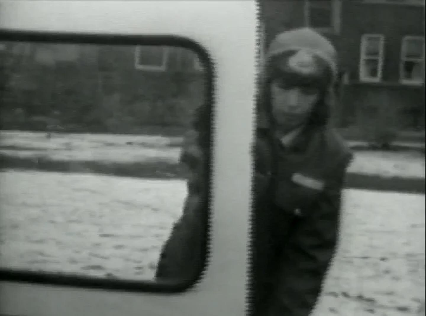 Black-and-white image of a young person standing beside a vehicle on a snowy street. The person wears a winter coat and a hat with ear flaps, partially obscured by the doorframe of the vehicle. The side of the vehicle dominates the foreground, while a house with multiple windows is visible in the background.