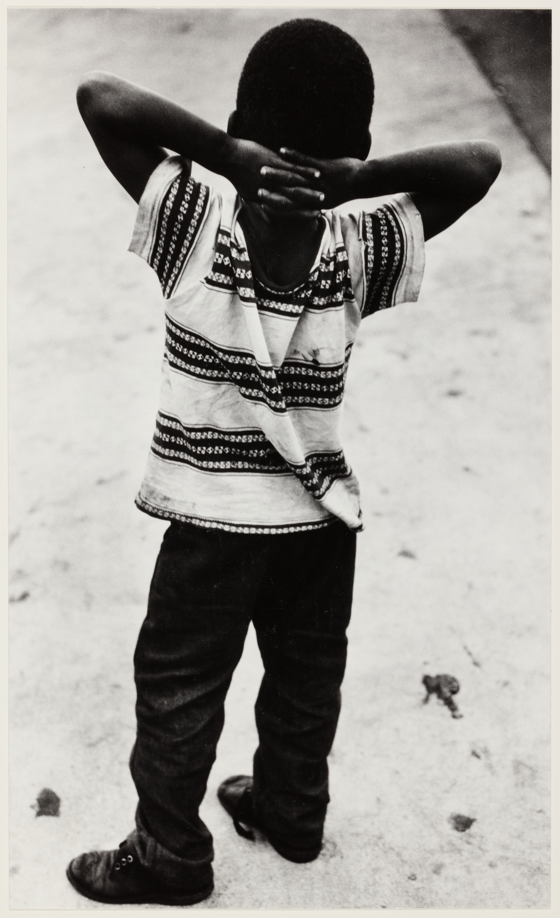 Black-and-white photograph of a young boy standing on a sidewalk with his back to the camera. His arms are raised, and his hands are clasped behind his head. He wears a patterned short-sleeved shirt with a tear down the back seam, dark pants, and worn shoes. The smooth pavement contrasts with the texture of his clothing.