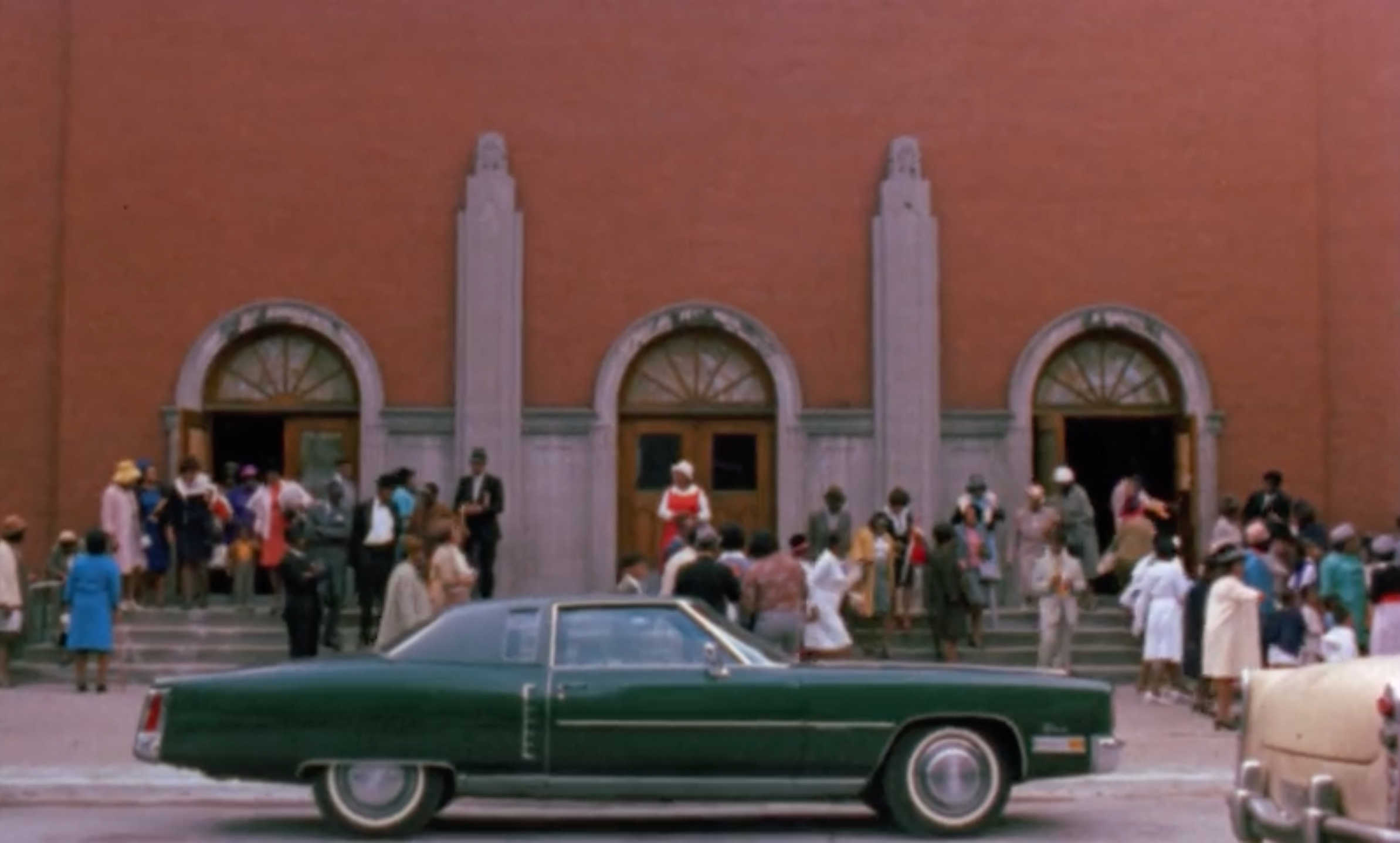 Crowd of people gathered outside a large brick building with three arched doorways framed in light stone. Most people are dressed in formal or semi-formal clothing, suggesting a special occasion. A dark green Cadillac-style car is parked prominently in the foreground along the curb, partially blocking the view of the building’s lower steps. Another light-colored car is visible at far right. The crowd appears to be socializing, entering, or exiting the building.